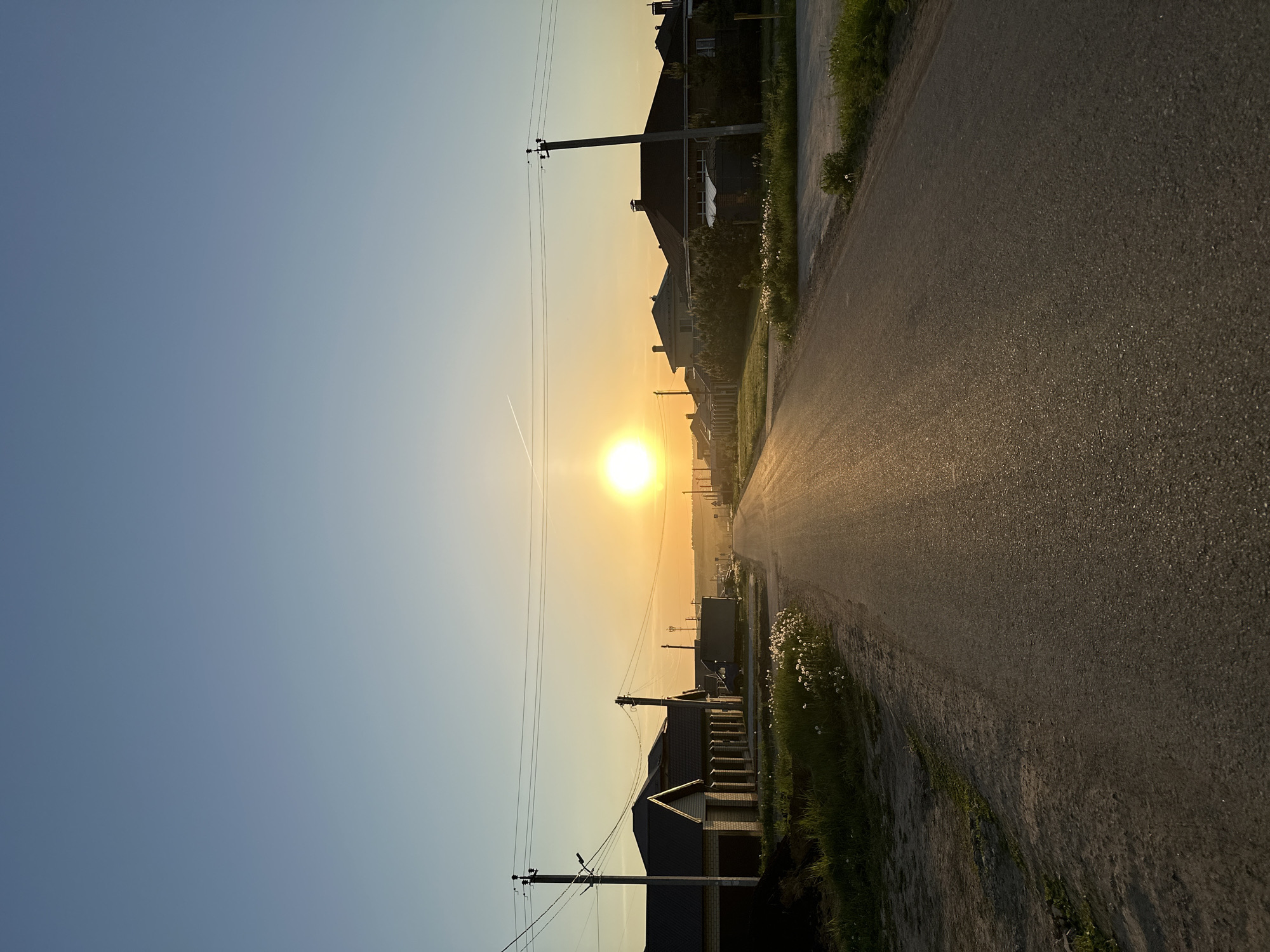 The image captures a street scene at sunset in Эстачи, Russia. A straight asphalt road recedes into the distance towards the setting sun, which dominates the horizon. On either side of the road are houses, visible as silhouettes against the bright sky. Utility poles with power lines run along the side of the street, adding vertical elements to the composition. The sky transitions from blue at the top of the image to orange near the horizon. There is a faint contrail from an airplane crossing the sky near the sun. Vegetation lines the edges of the road. The street appears wet, reflecting the light from the sun.