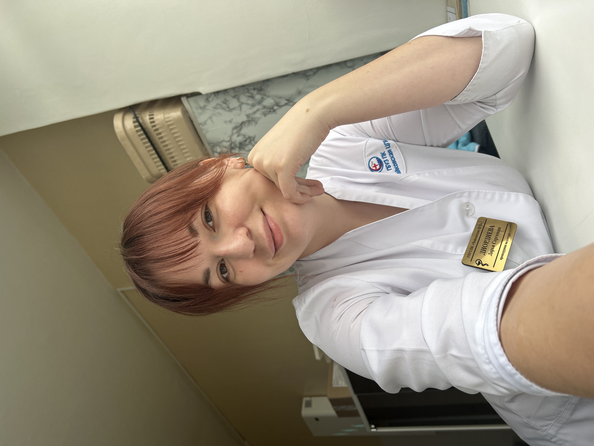 A fair-skinned woman with reddish-brown hair is seen from the chest up, angled towards the camera. She is wearing a white lab coat. A gold name tag with the name "DIORDIEVA Daria Sergeevna" in Cyrillic is pinned to her coat. The Russian words "procedure nurse" are visible on the tag. The upper left of the name tag reads "GBUZ PK 'Tchaikovsky TSGB' " also in Cyrillic, which when translated is the "Tchaikovsky Central City Hospital." The word "GBUZ PK" in Russian on the lab coat translates to "Healthcare Institution of the Perm Region." Her left arm is bent, with her hand touching her cheek. The background includes a light-colored wall and a partial view of a white surface that may be a table or counter. The location is near the city of Фоки in the Perm region of Russia.