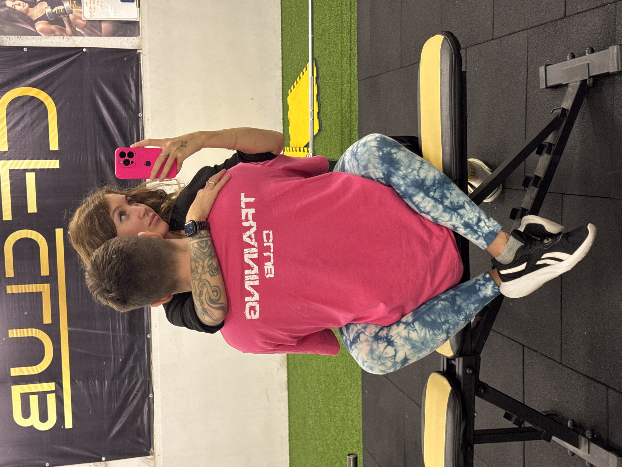 This image, taken in Chaikovsky, Russia, features two individuals in what appears to be a gym or fitness center. A woman with long brown hair, wearing a black top, is positioned behind and embracing a man with short brown hair. She holds a bright pink smartphone in her right hand, seemingly taking a selfie or video, and looks upwards and to her right. Visible details on the woman include rings on her fingers, a watch on her left wrist, and a tattoo on her right forearm. The man is wearing a pink t-shirt with the reversed text "TRAINING CLUB" across the back, blue tie-dye leggings, and black and white sneakers. A tattoo is also visible on his left arm.
The setting includes black rubber mat flooring and a strip of green artificial turf. To the right, a yellow and black exercise bench or machine is visible. On the left wall, a large black banner with yellow, reversed text clearly reads "ELECTRO CLUB". Above this banner, a poster shows images of a person lifting weights and some indistinct text. The overall scene suggests a casual and possibly affectionate interaction between the two, likely during or after a workout session.