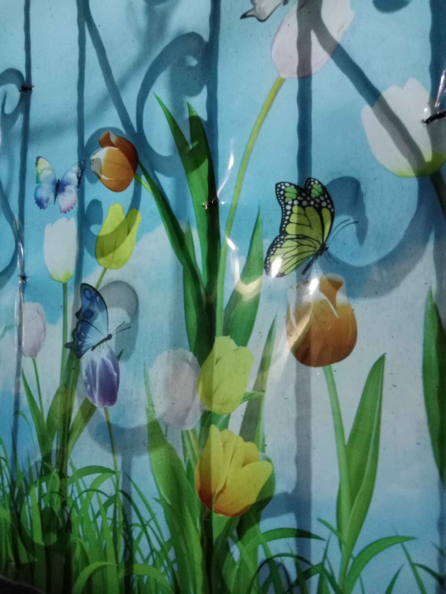 The image depicts a close-up view of a translucent, light blue curtain or banner adorned with a stylized floral and butterfly pattern. The pattern features several tulips in various colors including orange, yellow, white, and purple, interspersed with green leaves and grass. Three butterflies are visible: one light blue and pink, one predominantly blue, and one yellow with black markings. The background is a solid, textured light blue. The lighting suggests an indoor setting, potentially with ambient light casting shadows of vertical bars onto the patterned surface, creating a visual depth effect. The scene is devoid of any people or discernible landmarks of West Jakarta, Indonesia.