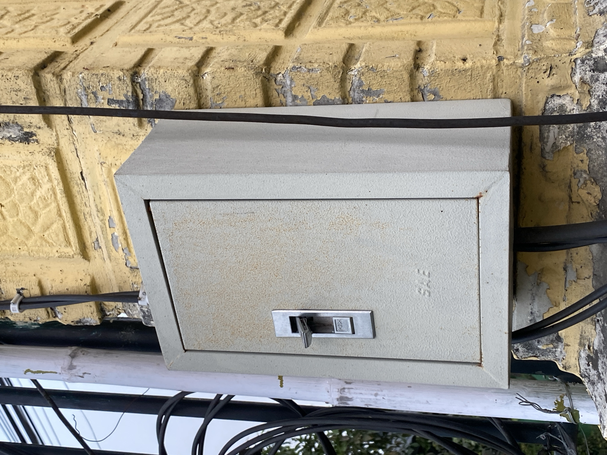 An outdoor electrical box is mounted on a distressed yellow brick wall in the city of Kasihan, Indonesia. The box is a light gray, rectangular enclosure with a door that has a silver-colored lever-style switch on the left side. The word "PUSH" is visible on the switch plate. Faded lettering that appears to be "SAE" is embossed on the front of the electrical box. The brick wall has areas of peeling yellow paint, revealing darker gray plaster and brick underneath. Several black electrical cables are visible, some running vertically along the wall and others entering the bottom of the electrical box. A white cylindrical pole is positioned to the left of the electrical box, also covered in some peeling material. Black cables are bundled and attached to this pole. The background shows a glimpse of green foliage and a wire fence.