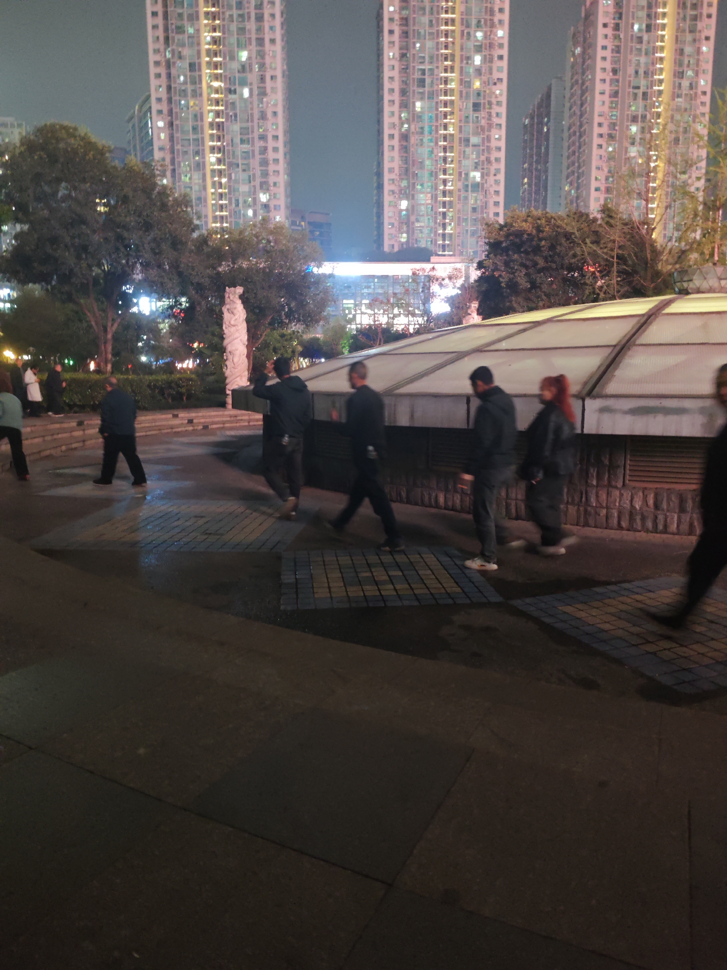 The image depicts a nighttime urban scene in Liangjiang, China. In the background, illuminated high-rise apartment buildings dominate the skyline. A group of people are walking through what appears to be a public square or park area. The ground is paved, with sections of brick or tile. A structure with a translucent roof is visible. To the left, there are trees, a statue, and streetlights. The overall lighting is dim, creating shadows and obscuring details.