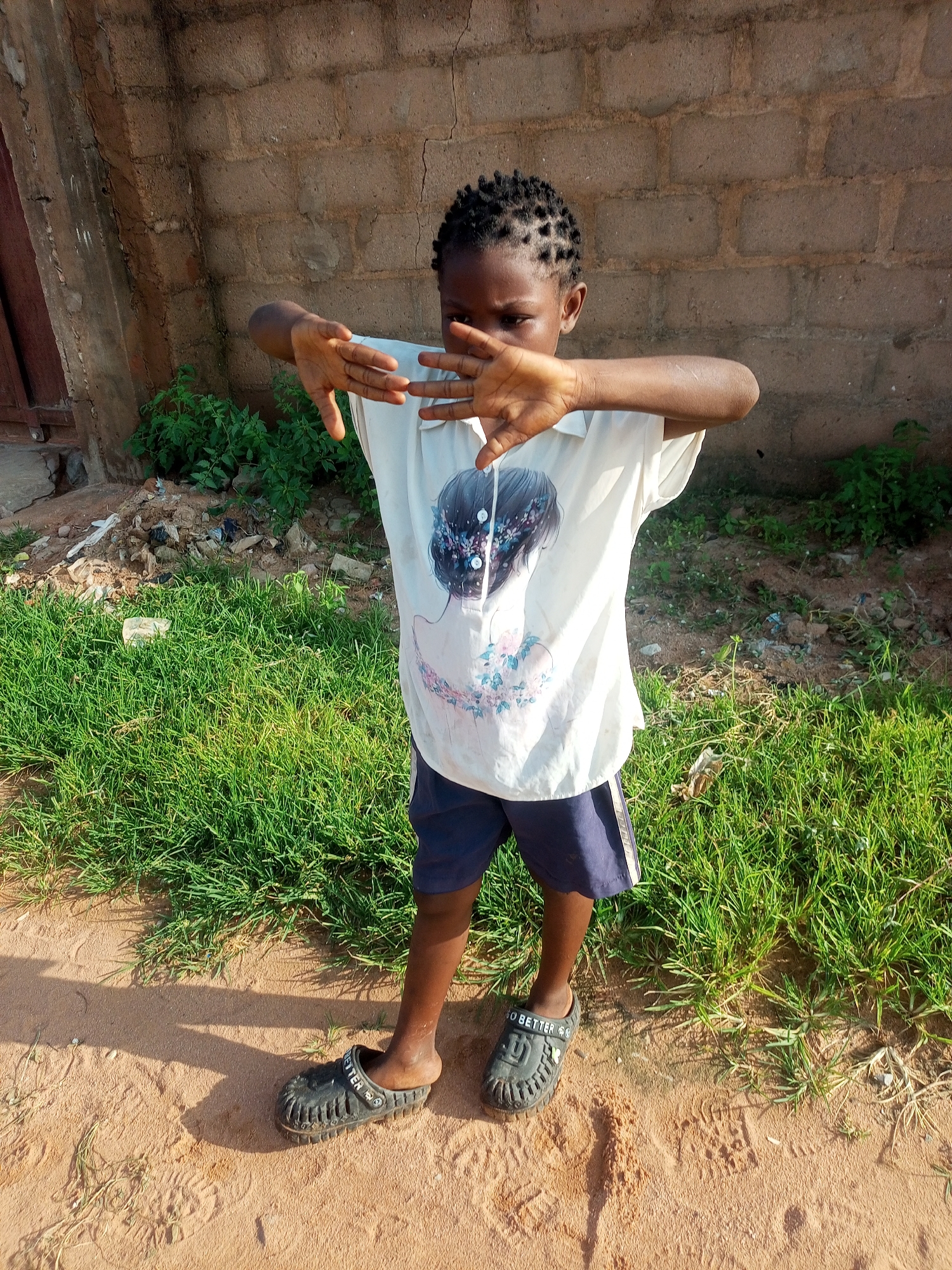 A young child stands in front of a brick wall and a patch of grass in Tempure, Nigeria. The child is wearing a white t-shirt with a graphic print of a person's back and a floral design, and dark blue shorts. On their feet, the child wears dark-colored slip-on sandals. The child's hands are held up in front of their face, obscuring it. The ground is sandy and scattered with dried leaves and small rocks. The brick wall appears weathered and has some cracks. The grass is green and somewhat unkempt. The scene is lit by natural sunlight, casting a shadow on the ground to the child's left.