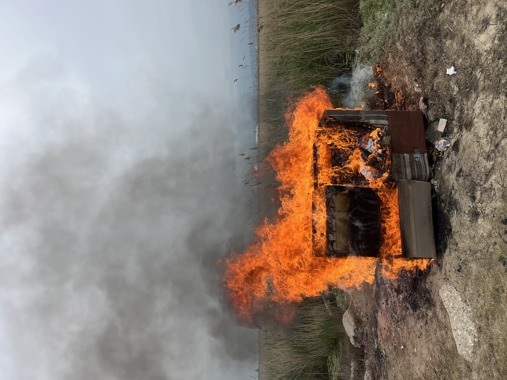 A burning couch stands on a patch of dirt and grass. Flames engulf the couch and thick grey smoke billows to the left. In the background, reeds grow along a flat, possibly marshy area under an overcast sky. The location is likely Kaspiysk, Russia.