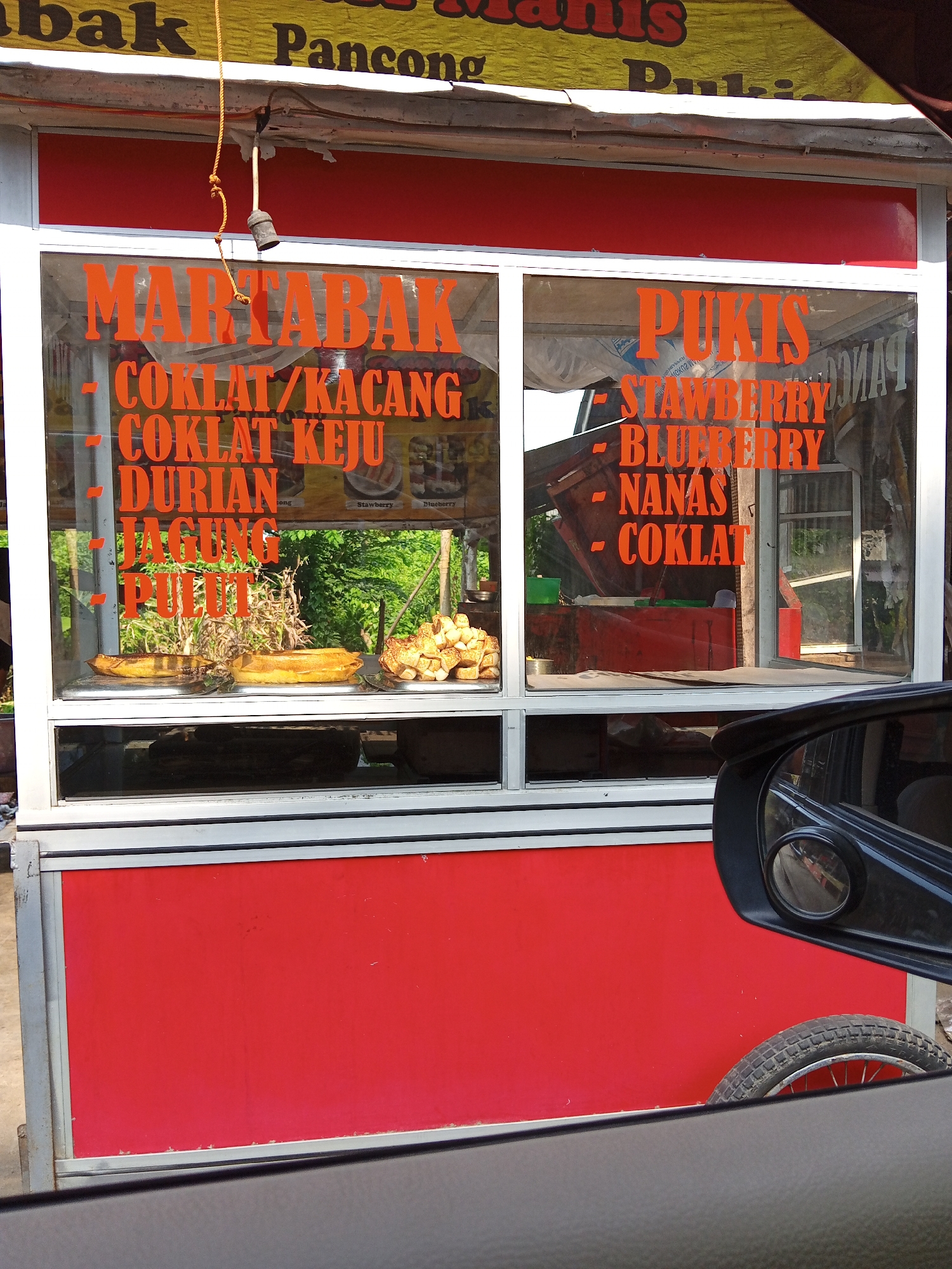The image shows a food stall in Keudesimpangempat, Indonesia, displaying signs for "Martabak" and "Pukis." The stall is red and white, with orange lettering on the glass windows listing flavors like "Coklat/Kacang" (chocolate/peanut), "Coklat Keju" (chocolate cheese), "Durian," "Jagung" (corn), and "Pulut" (glutinous rice) for Martabak, and "Strawberry," "Blueberry," "Nanas" (pineapple), and "Coklat" (chocolate) for Pukis. Food items are visible on the shelf behind the glass. The stall appears to be on wheels, with a portion of a wheel visible on the right. Part of a car, including the side mirror, is visible in the foreground. A yellow tarp is overhead. The backdrop shows green trees and foliage.