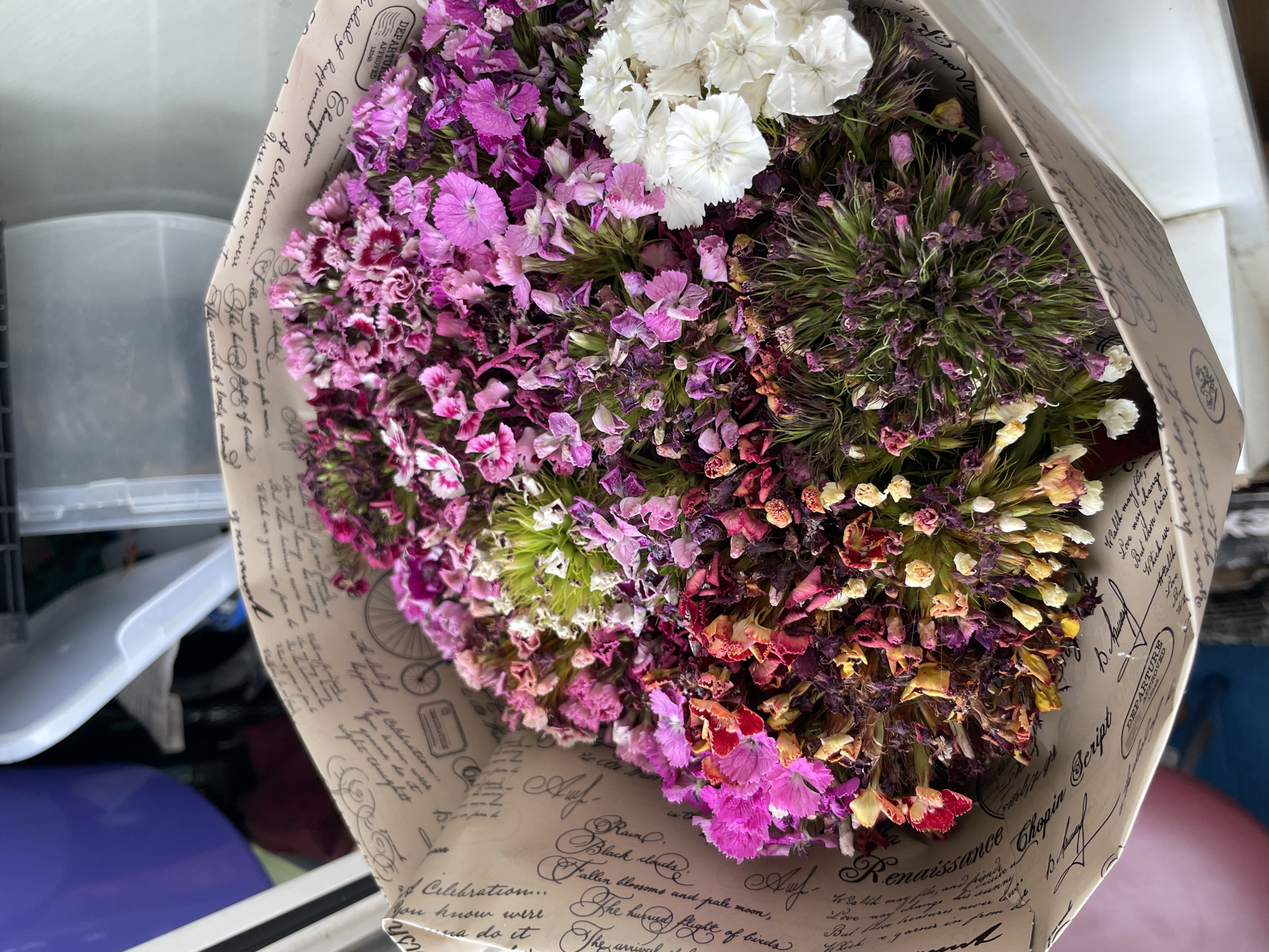 A bouquet of flowers, primarily in shades of pink and purple with some white and yellow accents, is presented in a decorative paper wrapping. The paper has a distressed, vintage look with handwritten script and graphic elements. The flowers themselves show signs of wilting, with some petals dried and curled. The background is out of focus but appears to be an indoor setting with storage containers visible. There is no indication of a specific location in Kyiv, Ukraine.