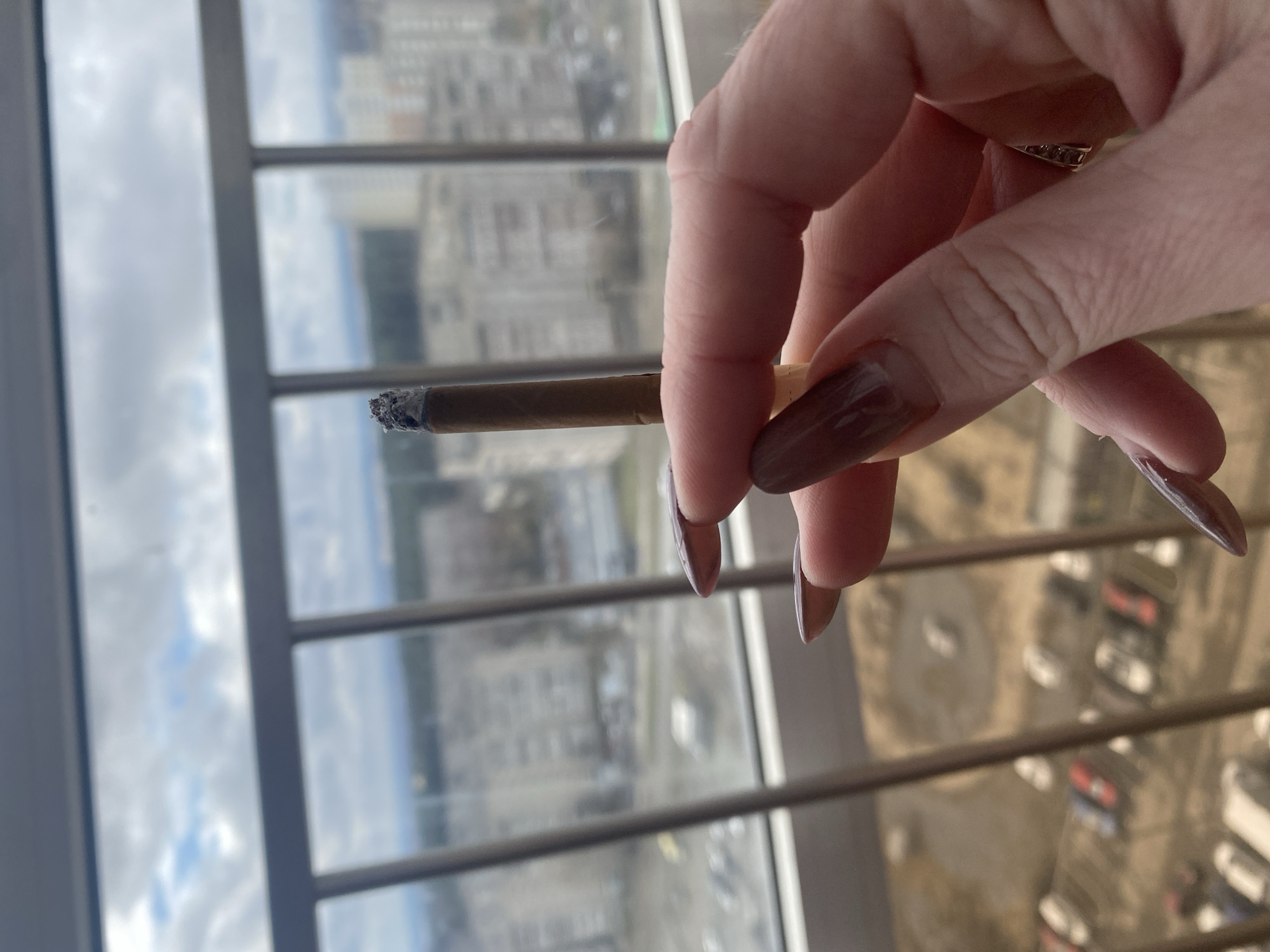 A hand holds a lit cigarette, appearing to be a hand-rolled one. The hand has long, brown-colored fingernails. The background shows a cityscape through a window. Based on the architectural style of the buildings visible through the window, this location is likely in Yekaterinburg, Russia. The buildings appear to be apartment complexes. The weather outside looks cloudy.