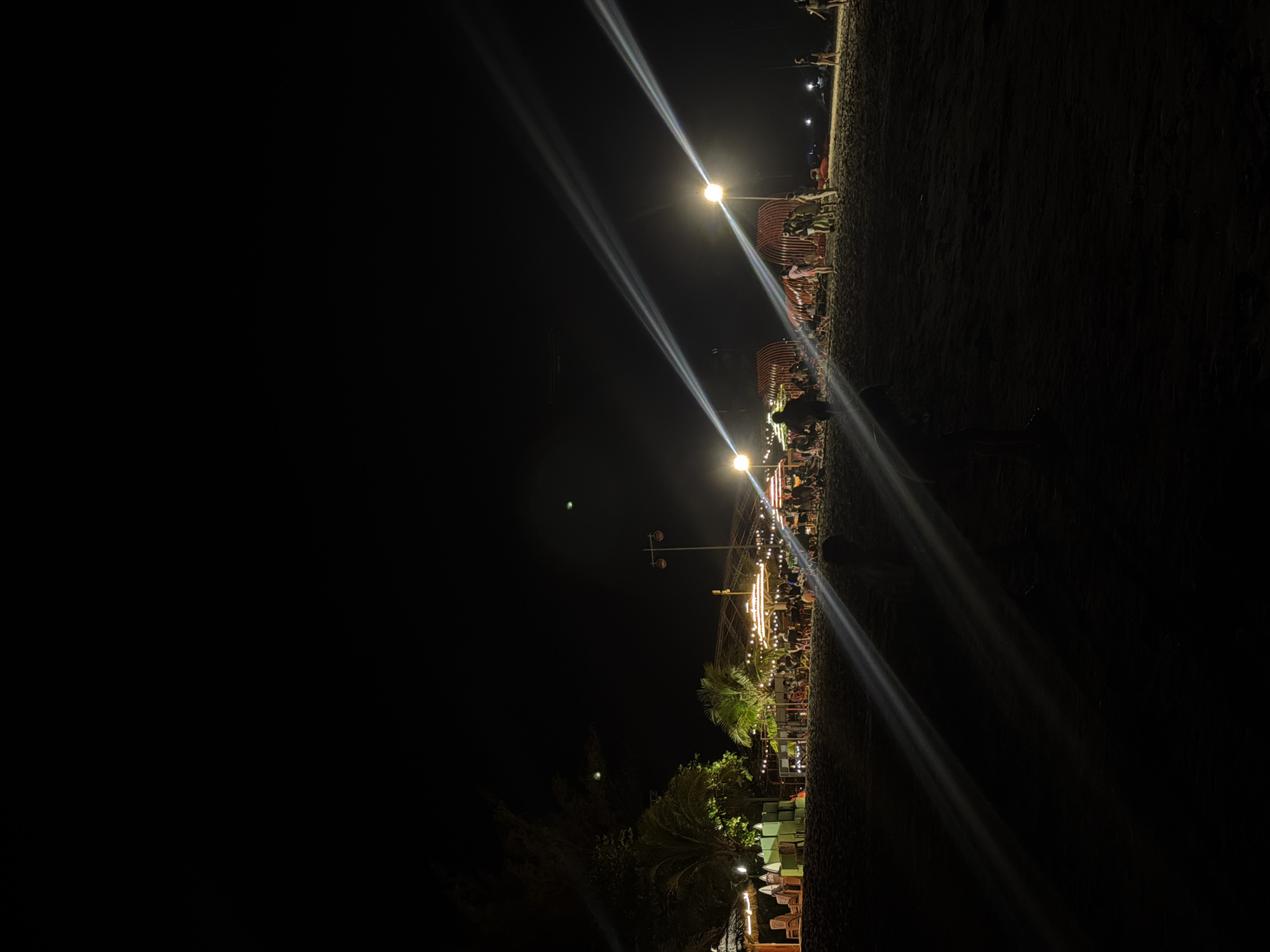 This vertical image, if rotated 90 degrees counter-clockwise, depicts an outdoor night scene of a bustling gathering or market. The upper half of the upright image is dominated by a dark night sky, while the lower half showcases a lively, illuminated area.
Numerous people are visible, mostly as silhouetted figures or partially lit individuals, engaged in various activities like walking, congregating, and sitting at tables. The setting includes a series of outdoor structures, possibly food stalls or seating areas, characterized by curved red frames and extensively decorated with string lights that cast a warm, festive glow. Several bright streetlights stand out, emitting intense, diagonal beams of light that cut across the dark expanse of the sky. These beams create prominent lens flares, adding to the visual dynamism.
Lush palm trees and other foliage are interspersed throughout the scene, particularly towards the lower left of the upright image, blending with the illuminated sections. The ground appears uneven and unpaved, suggesting a natural or semi-developed outdoor location. The overall atmosphere is one of a vibrant nocturnal event. The time of day is clearly night, with all illumination coming from artificial sources. No discernible text is visible within the image. The scene conveys a sense of community and leisure, consistent with an outdoor market or social gathering in a location like Dong Hai Ward, Vietnam.