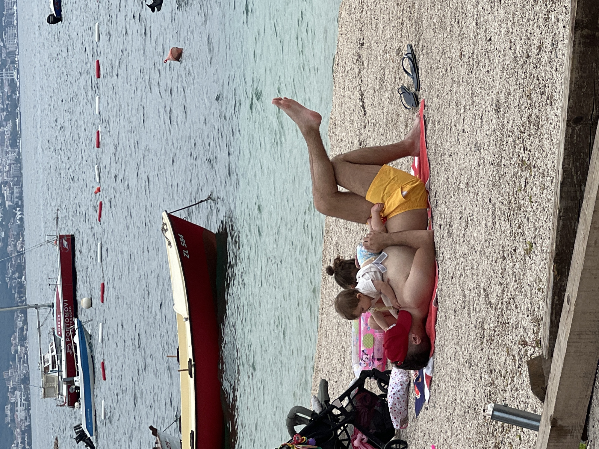 The image depicts a tranquil beach scene in Baošići, Montenegro, during daylight hours. In the foreground, an adult male is lying on his back on a red beach mat on a pebbly shore, wearing yellow swim shorts. His legs are bent, with one foot raised in the air. Two young children are interacting with him, resting on his chest and stomach; one child wears a red hat. A pair of black flip-flops are placed near the adult's head, and a black baby stroller, along with other belongings like towels and blankets, is partially visible to the left.
The beach meets calm, light blue water. Several boats are present in the water. Closest to the shore is a red and cream-colored boat with the registration "ZL 584" visible on its stern. Further out, a larger red and white boat displays the text "PORTONOVI MONTENEGRO" on its side. A line of red and white buoys stretches across the water, likely marking a swimming area. A white spherical buoy and a small, reddish-brown floating object are also visible in the sea. In the distant background, a town or cityscape extends along the coastline, backed by hills. The scene conveys a relaxed and leisurely family moment by the sea.