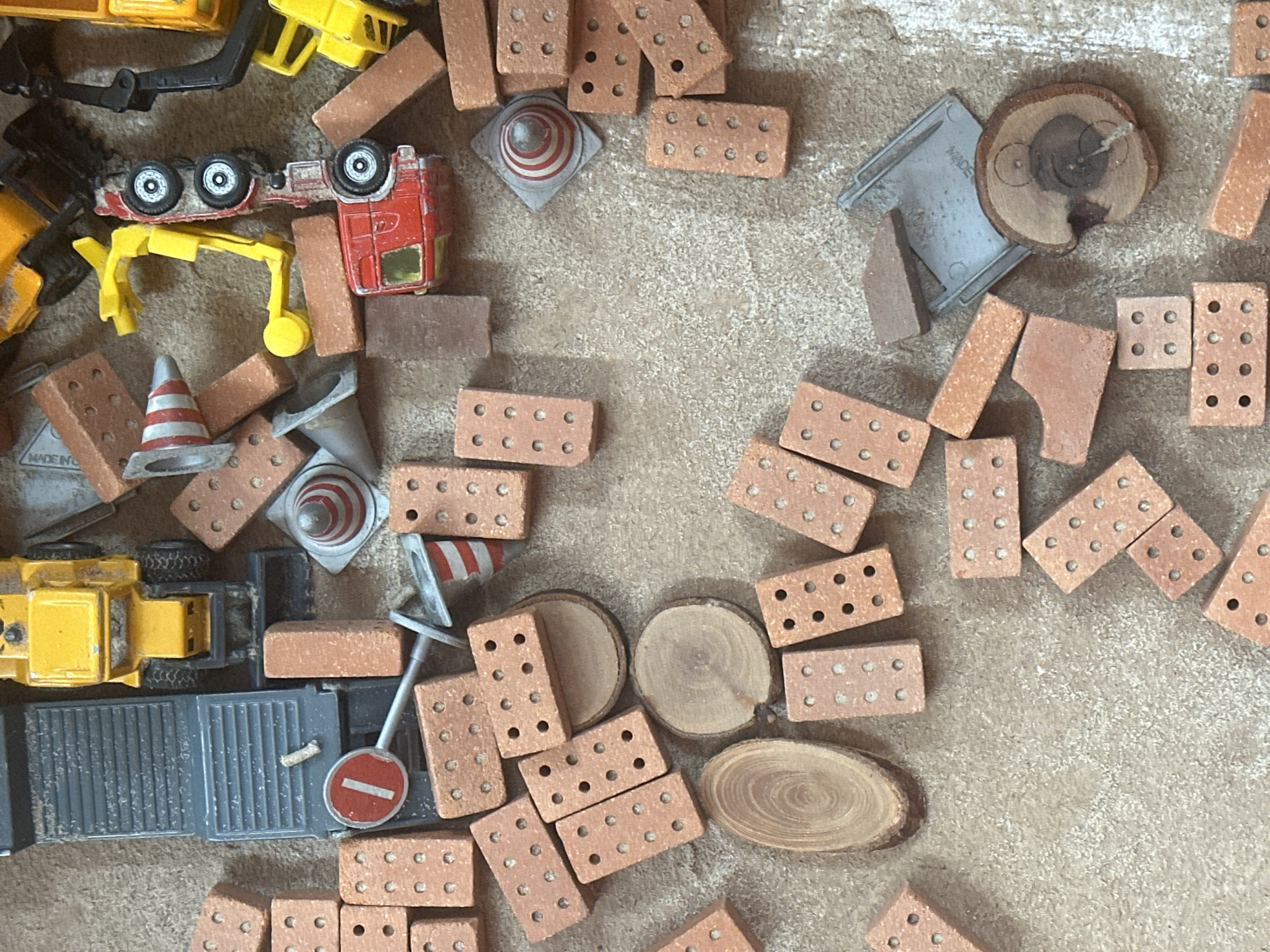 This is a top-down, close-up shot of a collection of toy construction items scattered on a textured, light brown surface that resembles sand or sawdust. The subjects are primarily toy construction elements, including miniature bricks, construction cones, a toy truck with a trailer, and parts of toy construction vehicles. There are also several small, circular wooden discs, possibly intended as building materials.
The scene depicts a disorganized pile of these toys, suggesting a play area or a scene from a child's imaginative construction project. There is no discernible activity or event taking place; the toys are simply arranged.
Notable details include the variety of toy bricks, some with visible "holes" or indentations. The toy truck is a prominent object, with a yellow cab and a grey trailer. The construction cones are striped red and white. The wooden discs have visible wood grain.
No people are visible. The lighting suggests an indoor setting, possibly with artificial light, as there are no strong shadows indicating natural sunlight. The time of day and weather conditions are not determinable. The location is unknown, but the material the toys are on could suggest a sandbox or a craft area.
There is some faint text visible on the grey metal piece underneath one of the wooden discs, which appears to say "MADE IN". The text is difficult to read clearly.