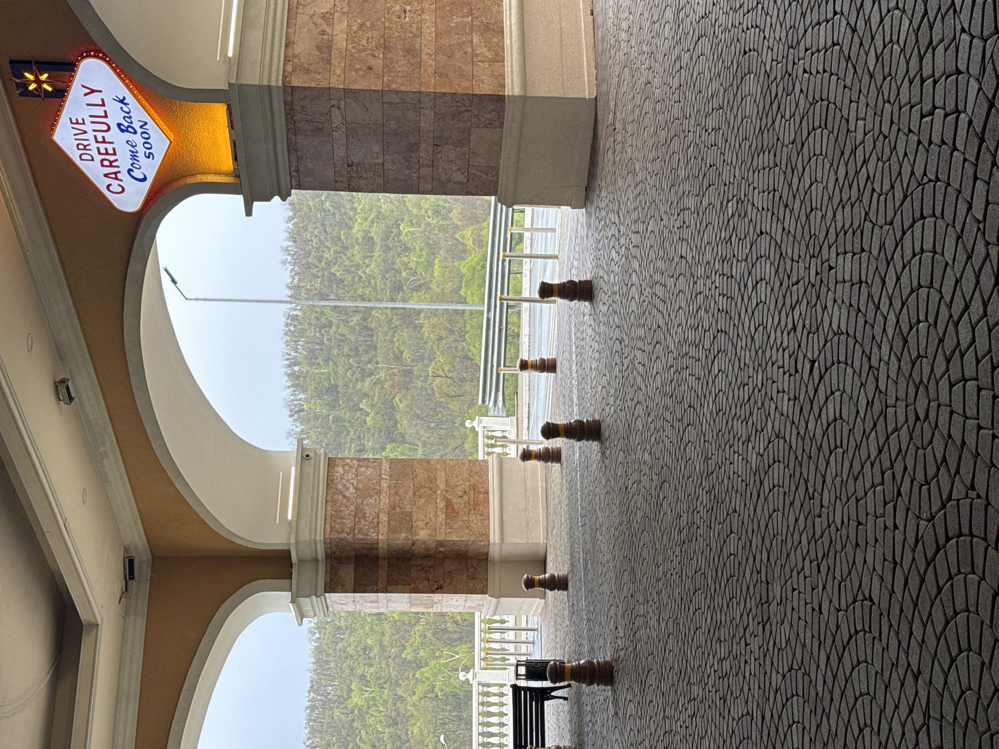 The image depicts a covered walkway with decorative columns and arches, leading towards a roadway with a metal barrier and a distant view of greenery, possibly trees. The walkway is paved with a dark gray cobblestone pattern in an arched design. The columns are clad in marble with beige or cream-colored accents, and the arches are finished in a similar light tone.
Along the walkway are a series of dark brown bollards with yellow stripes. A black bench is visible in the lower portion of the image, between two of the bollards.
To the left of the frame is a vintage-style sign with the words "DRIVE CAREFULLY Come Back SOON" in a classic font, indicating a place or destination. A starburst emblem is above the text.
The overall scene suggests a location with architectural features and a pathway leading out of an enclosed area towards a road or highway. The setting could be Shkotovo, Russia.