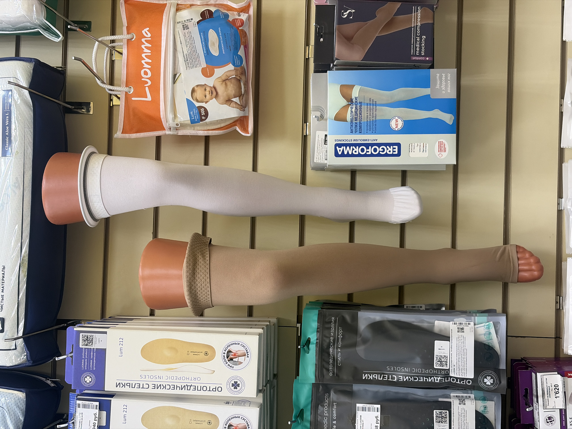 This is an indoor scene showcasing a store shelf likely in Saint Petersburg, Russia, filled with orthopedic and medical supplies.
The shelf features mannequins wearing compression stockings. There are packages of "Luomma" orthopedic insoles and anti-embolism stockings ("Ergoforma"), along with products from other brands that include "Classic Aloe Vera L" mattresses, a baby product in orange packaging labeled "Luomma," and medical compression stockings from an unreadable brand in black packaging.
The scene is well-lit and provides a clear view of the products on display.