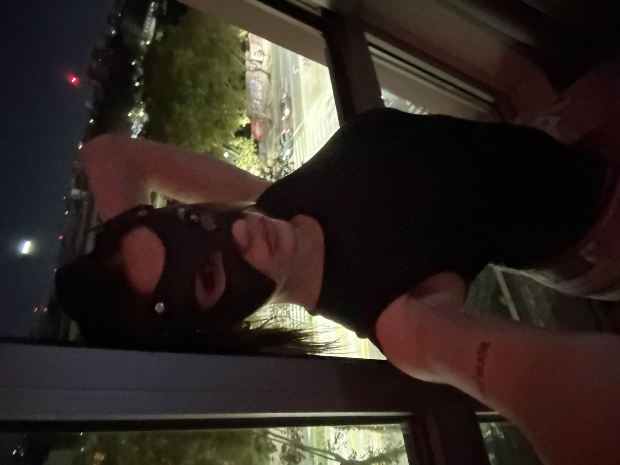 The image shows a person positioned indoors by a window, looking out over a city at night. The person is wearing a black short-sleeved top and a black mask that covers their face, resembling a pup hood, with dark hair visible. Their arms are raised, resting against the window frame or glass.
The scene outside the window is an urban environment illuminated by city lights, indicating it is nighttime. A dark sky is visible, along with distant red lights and numerous white lights from buildings and streets below. Green foliage and other structures can be seen in the background, suggesting an elevated viewpoint over a city like Buenos Aires, Argentina.
On the person's right forearm, near the wrist, a faint reddish tattoo or marking is visible, which appears to spell "PUPPY". The lighting is dim, with the person partially silhouetted against the brighter city lights. The overall atmosphere is enigmatic due to the mask and the nocturnal setting.