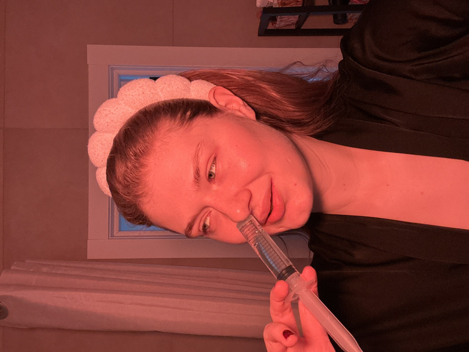 An adult female is observed in an interior room located in Nikol'skii, Russia. She is performing a nasal irrigation procedure, holding a clear plastic nasal irrigator with a plunger to her right nostril. The device contains a clear liquid with blue volume markings. The woman has light skin, brown hair styled away from her face, and wears a light-colored, textured headband. She is dressed in a dark, possibly black, satin or silk robe. Her left hand, featuring red nail polish on the thumb, grips the irrigator. Her gaze is directed towards the upper right, and her mouth is slightly open. The scene is illuminated by a prominent reddish-orange light. The background includes light-colored walls, a white framed doorway or opening with a blue hue visible through it, a white curtain on the left side, and a dark shelving unit holding baskets on the right.
