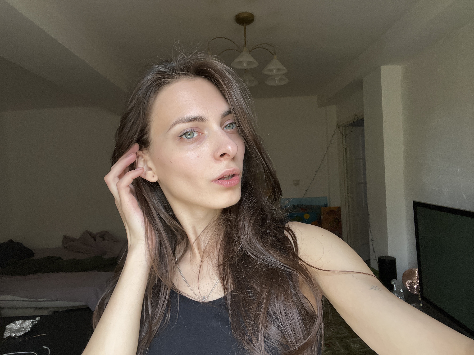 A medium, eye-level, selfie shot captures a fair-skinned woman in an indoor setting. The woman has long brown hair and fair skin. She has on a black tank top and a silver necklace. Her left hand is at her ear. The background reveals an interior scene with light-colored walls, a chandelier-style ceiling light, an unmade bed, a television set, and an abstract painting. While a general sense of an interior location is conveyed, there is no information in the image to suggest the city of Krasnoyarsk, Russia. It is not possible to infer the specific location or city from the image alone.