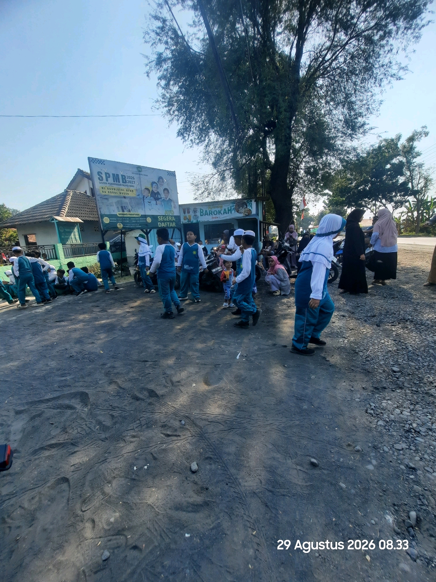 The image depicts a street scene in Sukokerto, Indonesia, with a focus on a group of children wearing school uniforms. The children, primarily boys, are dressed in teal and white attire, with some wearing white caps. Several are seated on the ground, while others are standing or walking. In the background, a building with a green facade and a large banner displaying "SPMB 2026-2027" is visible. The banner also features images of children and text in Indonesian. Adjacent to this is a small shop or stall with the word "BAROKAH" visible. Adult figures, mostly women in hijabs, are present in the background, some appearing to be accompanying the children. The ground is covered in dark, granular material, possibly ash or dark soil, with tire tracks and footprints visible. The environment appears to be an outdoor setting, with a large tree casting shadows. The sky is clear and blue, suggesting daytime. The lower right corner of the image shows a timestamp: "29 Agustus 2026 08.33".