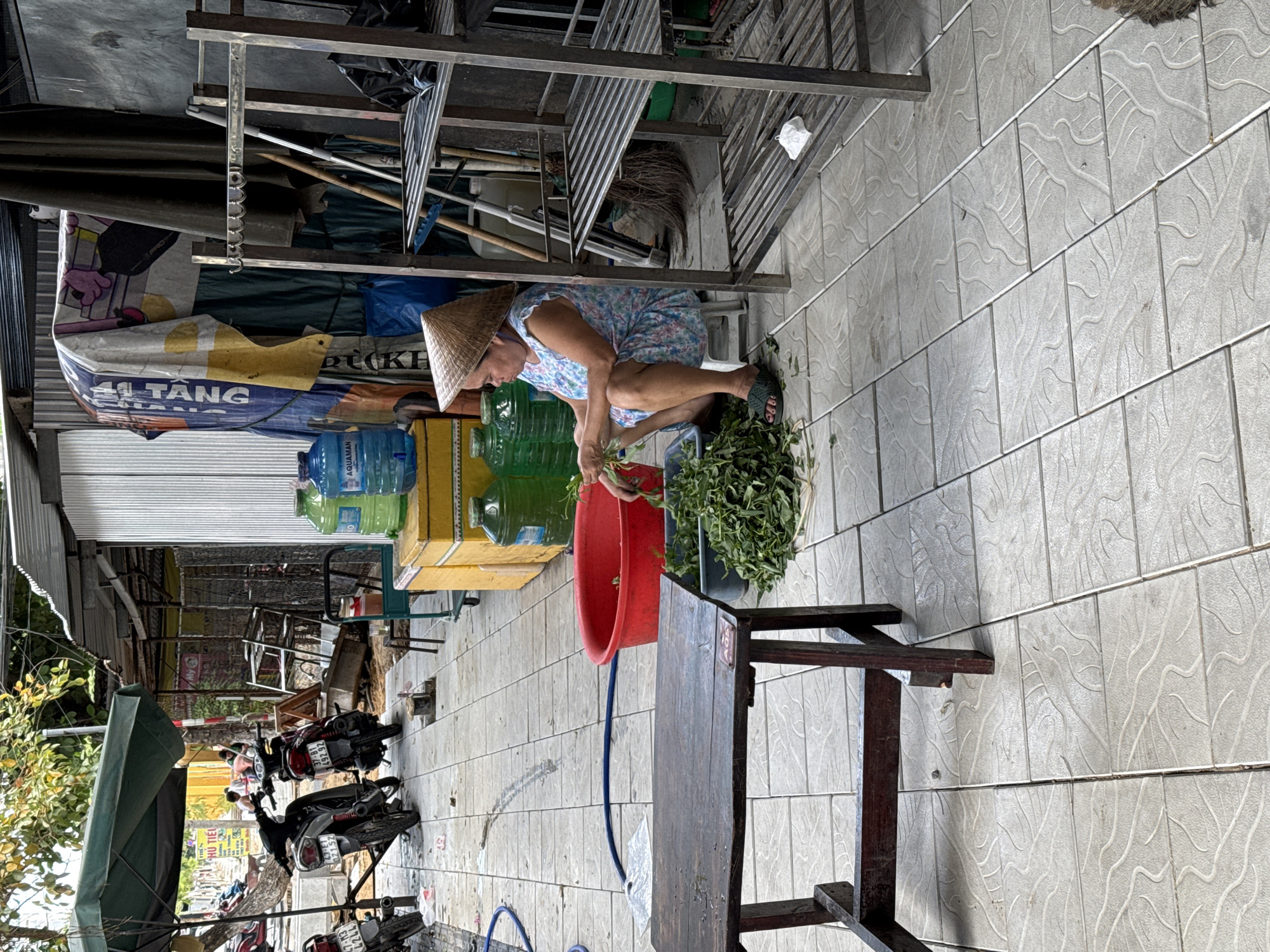 A woman wearing a conical hat is squatting on a tiled patio, preparing a large bunch of leafy green vegetables. She is dressed in a floral-patterned shirt and blue shorts, and is wearing dark sandals. To her right, a red plastic tub sits on the ground, and next to that is a large pile of the prepared greens. Behind her, stacked items include large plastic water bottles, one labeled "AQUAMAN," and yellow boxes. Further in the background, a metal shelving unit is visible, holding various items including what appears to be cleaning tools and bags. In front of the shelving unit, a colorful tarp is draped, with visible text including "TANG" and what looks like Vietnamese characters. The scene appears to be outdoors, possibly in a market or a roadside stall, given the presence of the shelving and stacked goods. To the left of the frame, several motorcycles are parked, and a green umbrella canopy is visible. The tiling on the ground has a textured, wave-like pattern. The overall impression is of a busy, everyday scene in a Vietnamese setting, likely during daylight hours.