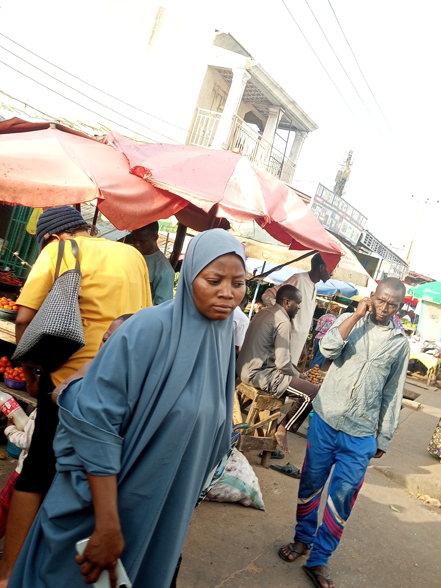 The image depicts a bustling outdoor market scene, likely in a warm climate given the attire and bright sunlight, suggesting midday. Several people are present, interacting within the market environment.
In the foreground, a woman wearing a light blue hijab and a long, flowing blue garment is walking towards the viewer, holding a silver smartphone in her right hand. Her expression appears serious. To her left and slightly behind her, a person in a yellow t-shirt and a dark blue beanie is visible from the back, carrying a patterned tote bag over their shoulder. Another individual in a grey t-shirt is seated on a wooden stool, looking downwards. A man in a light blue patterned shirt and blue track pants with red and white stripes is walking away from the viewer, holding a phone to his ear. He is wearing dark sandals.
Behind the main subjects, there are market stalls with red and pink umbrellas providing shade. Tomatoes are visible in bowls at one of the stalls. In the background, more people and stalls are present, along with a building with an ornate balcony and decorative columns. A sign with text is partially visible on a building in the mid-background, reading "UB MODERN CO" followed by illegible characters, possibly indicating a business or shop. The overall atmosphere is that of an active marketplace.