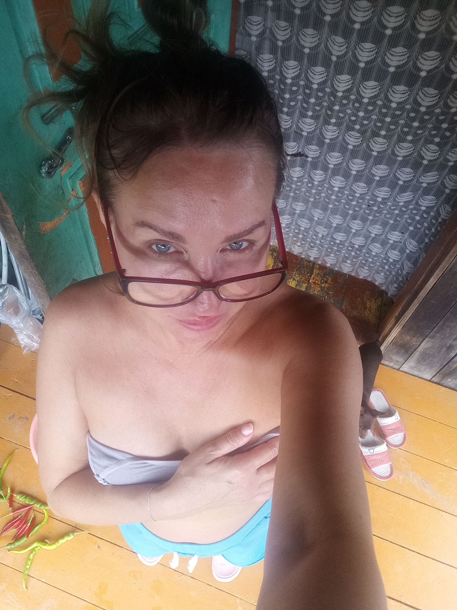 A woman with dark hair pulled back and wearing red-rimmed glasses is looking at the camera. She has one hand on her chest, covering it. She is wearing a light grey strapless top and light blue shorts with a white trim. In the foreground, there are several chili peppers on a wooden floor. In the background, there is a wall with a green and orange pattern and a patterned curtain. A pair of pink and white striped slippers are visible on the floor to the right of the woman.