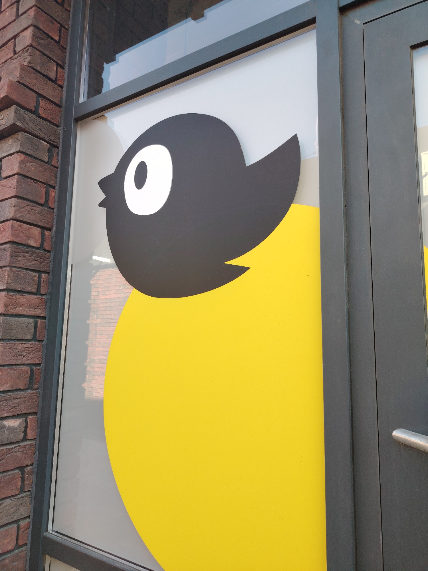 The image shows a close-up of a window with a large graphic of a stylized bird. The bird has a black head with a large white eye and a yellow body. The window is framed by dark gray metal and a brick wall is visible to the left. The graphic appears to be a decoration or branding for a business located in Rostov-on-Don, Russia. The scene is outdoors, and the lighting suggests it is daytime. There are no people visible in the image. The overall impression is a clean and modern storefront design.