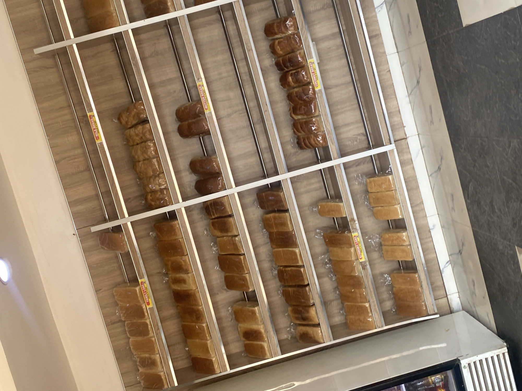 The image displays an interior scene of a retail establishment, likely a bakery or bread store, located in Rido, Nigeria. The central focus is a multi-tiered display unit constructed from light wood-grain paneling with horizontal silver metal rails. The shelves are densely stocked with numerous loaves of bread, each individually wrapped in clear plastic packaging. Visible labels on different shelves identify bread types, including "FAMILY LOAF," "MILK BREAD," "BUTTER BREAD," "COCONUT BREAD," and "ECONOMY LOAF." To the immediate left, a portion of a white commercial refrigerator or display cooler is visible, containing bottled items. The floor is tiled with large dark grey sections, complemented by a narrow strip of white and grey marbled tiles running along the base of the shelving unit. The ceiling is a light, neutral color and features a single illuminated recessed light fixture in the upper left quadrant of the frame. No individuals, actions, or interactions are present in the scene.
