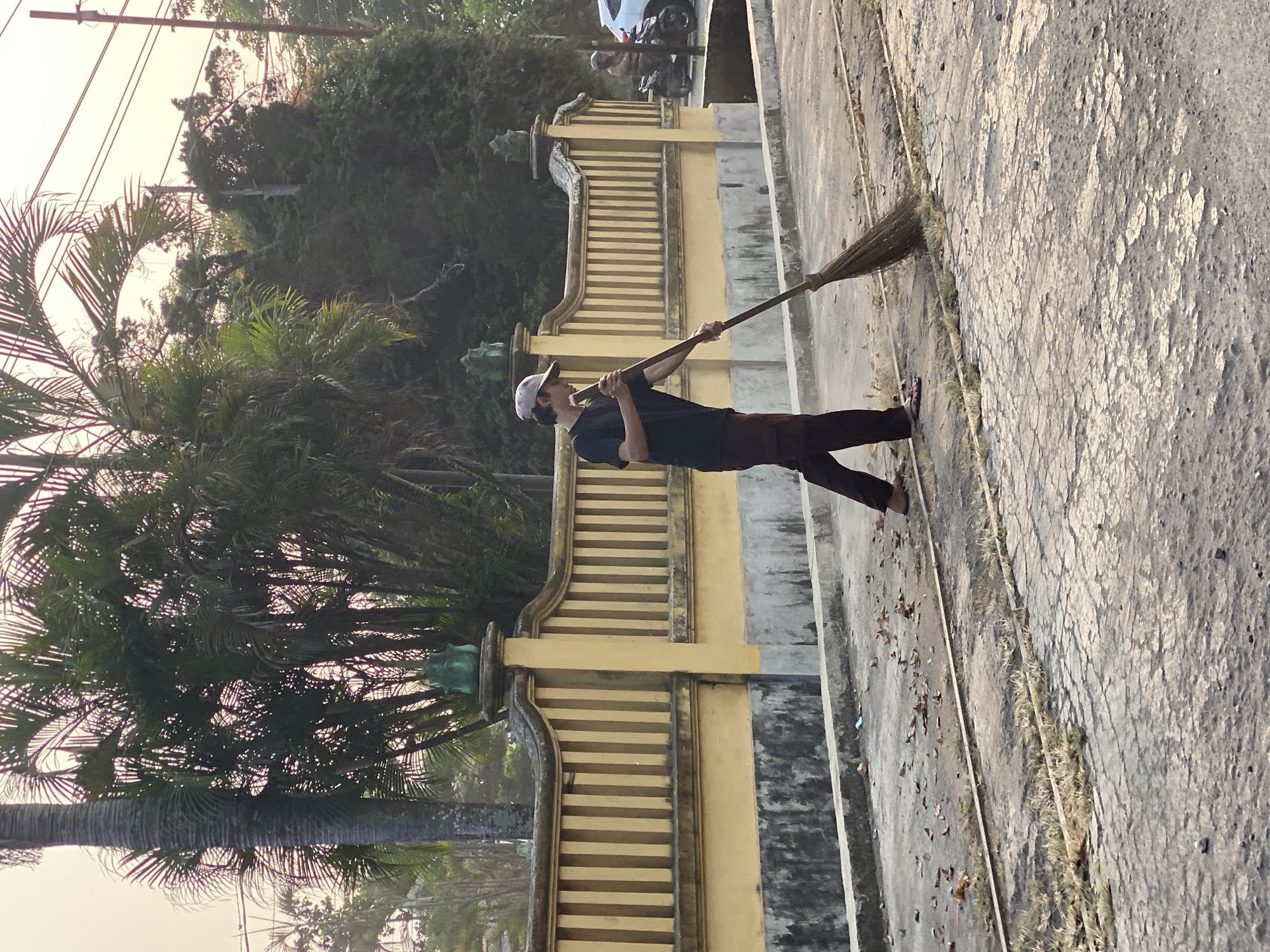 A young man wearing a dark t-shirt, brown pants, and a baseball cap is sweeping the ground with a broom. He is standing on a cracked concrete surface next to a low, yellow wall with vertical bars. Behind the wall, there are palm trees and other foliage. The scene appears to be outdoors, possibly in a residential area or along a street. A motorcycle with a rider is visible in the background on the right side of the image, and a white car is parked behind it. The lighting suggests it is either early morning or late afternoon. The location is identified as Kasihan, Indonesia.