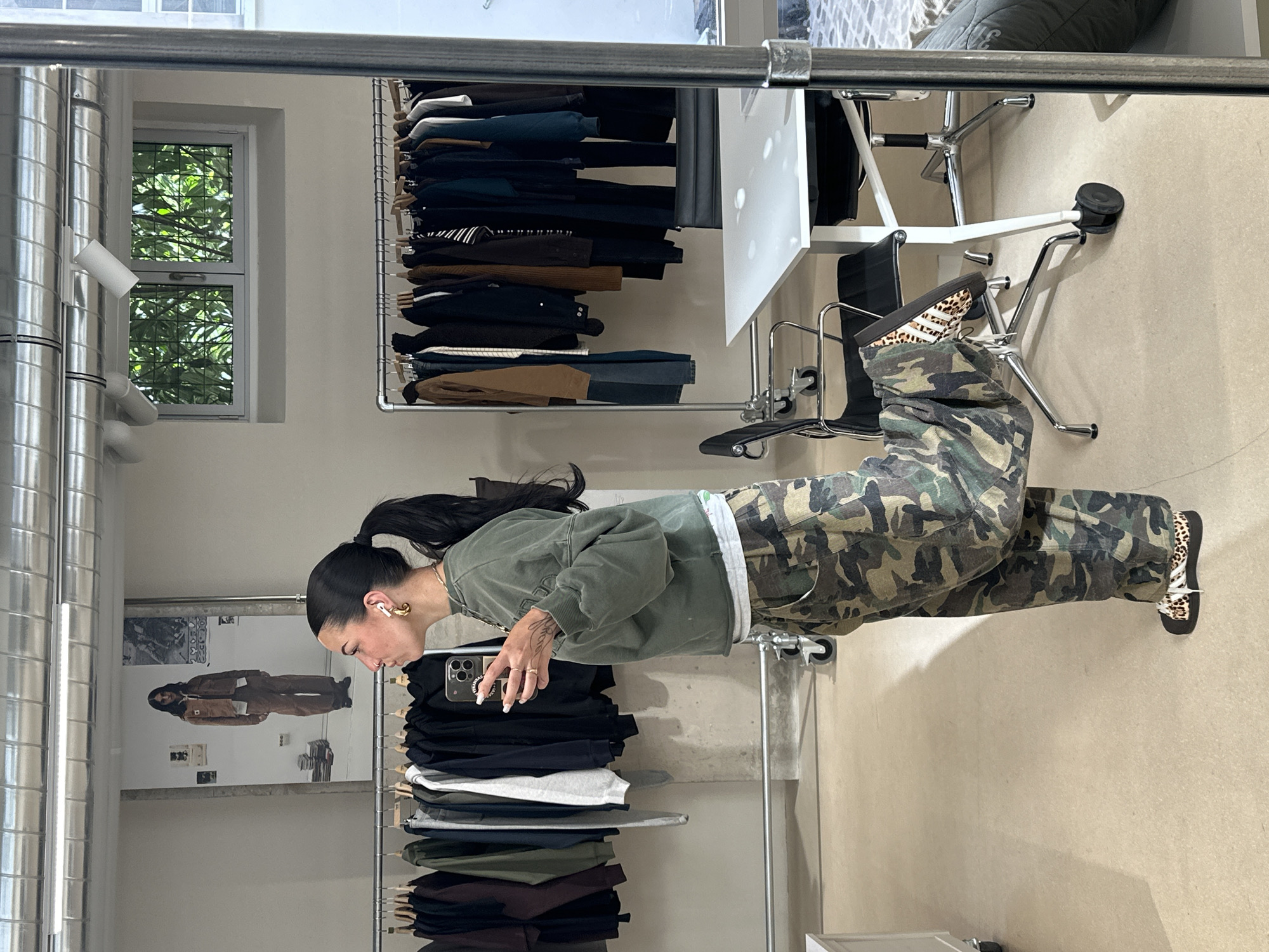 The image shows a reflection in a large mirror, capturing a scene inside a clothing store.
A woman stands in the center, facing the right side of the frame. She wears a green sweatshirt, camouflage pants, and leopard-print shoes. She holds a phone in her hand.
A metal clothing rack is situated behind her, holding folded clothing in various colors, including black, grey, and brown. Another clothing rack with hanging clothes is on the opposite side.
A white table and chairs are behind the woman.
A window is visible, suggesting an outside view of trees, and a poster with a person in brown clothing hanging on the wall.
The floor is a plain beige color.
This scene most likely takes place in Paris, France. However, I can't specify the exact location in the city.