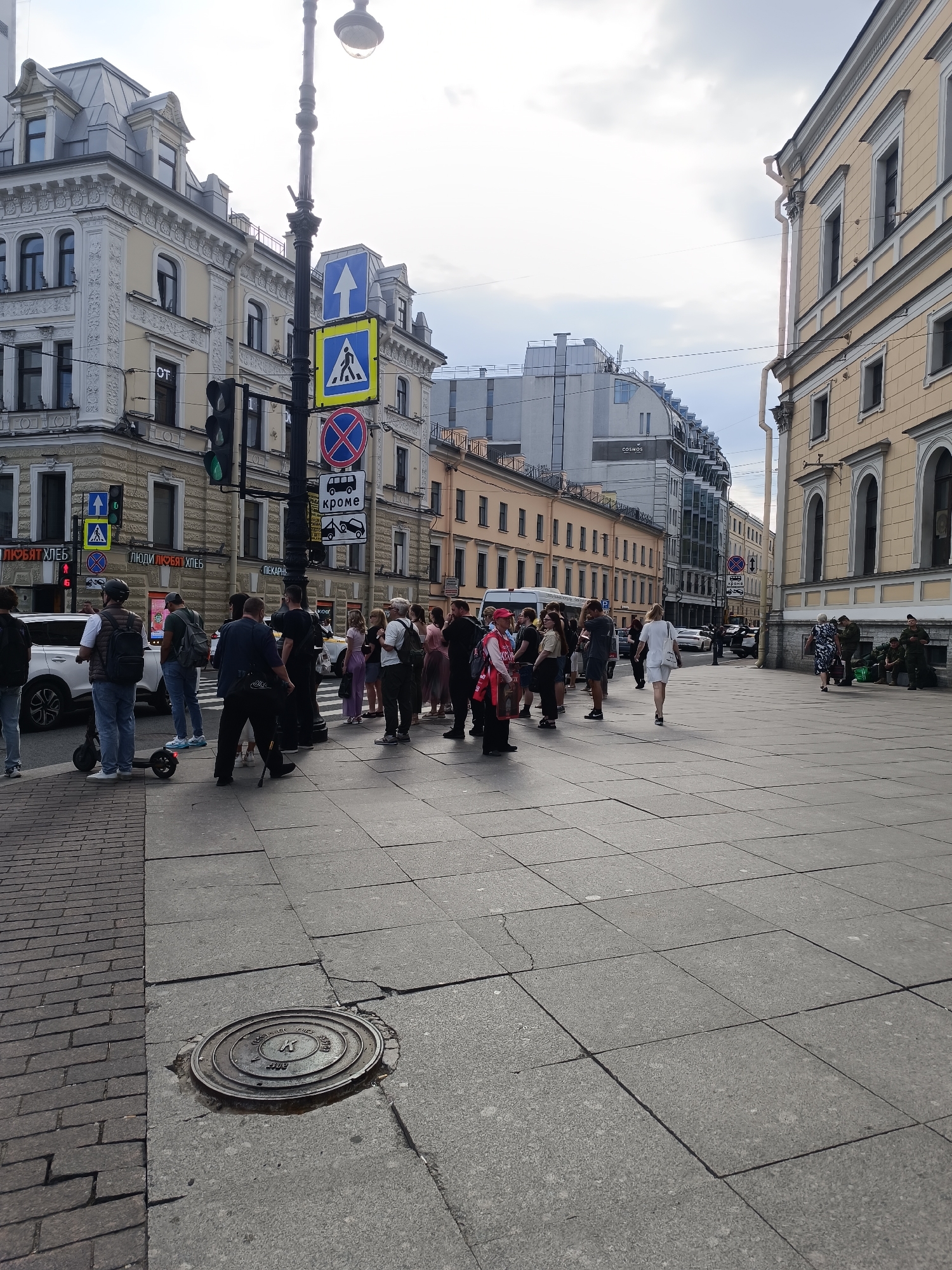 A street scene in Saint Petersburg, Russia, shows a group of people gathered at a crosswalk near a traffic light and several street signs, including pedestrian crossing signs and a no parking sign. In the background, ornate buildings line the street, with a modern glass building visible further down. Several cars, including a white SUV and a white van, are present, along with an electric scooter. The pavement is paved with large stone tiles, and a manhole cover is visible in the foreground. The sky is overcast.