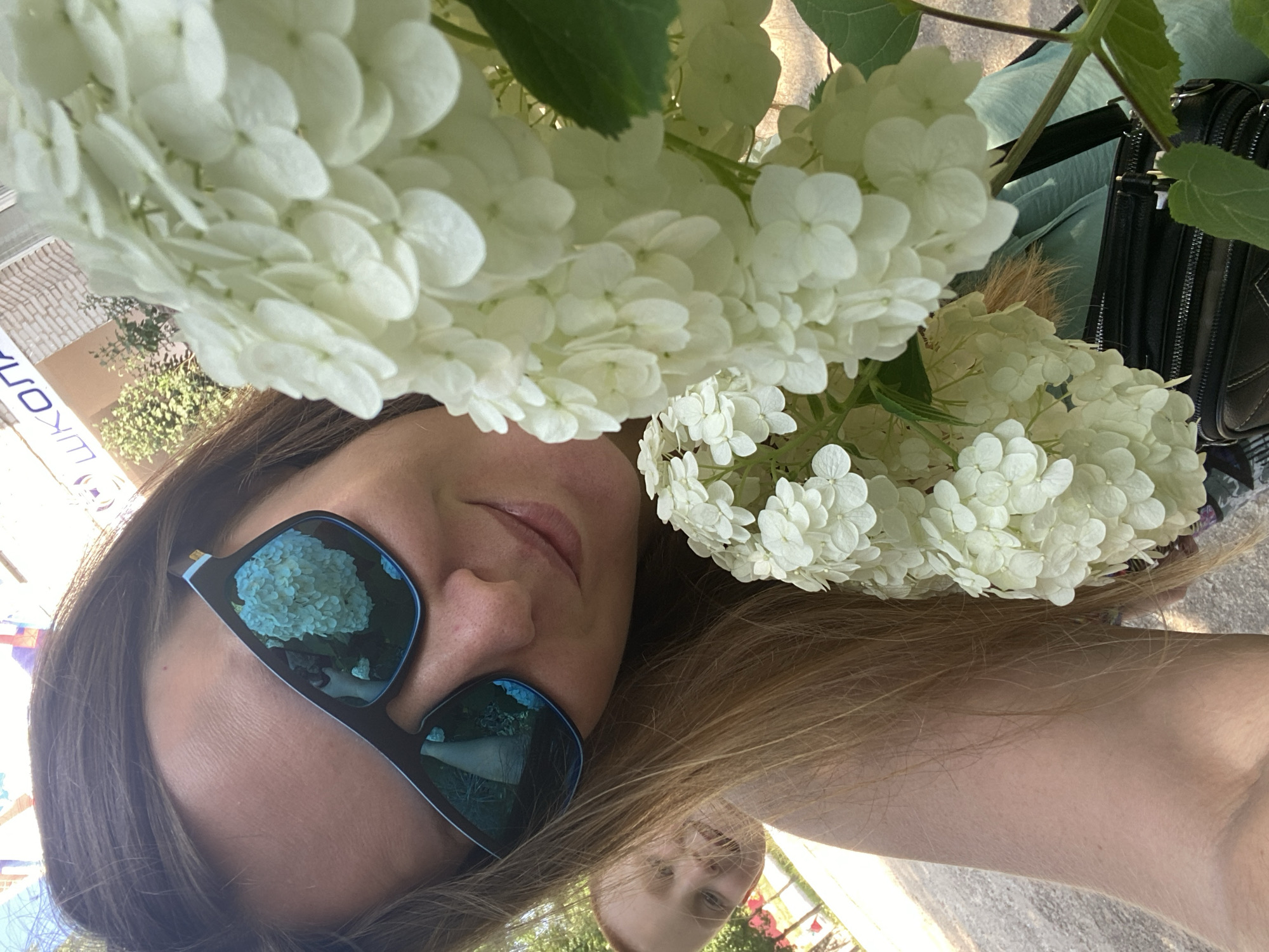 In this close-up selfie, a woman with long brown hair, wearing dark sunglasses with blue rims, smiles while holding a large bouquet of white hydrangeas. In the background, slightly out of focus and to the left, a young child with red hair and fair skin peeks out from behind the woman's shoulder, also smiling. The woman is outdoors, and the background suggests a street or public area with a building that has signage, possibly a school, with Cyrillic lettering reading "ШКОЛА" (SHKOLA, meaning school). The overall scene is bright and appears to be taken on a sunny day, likely in the city of Noviy, Russia, given the signage.