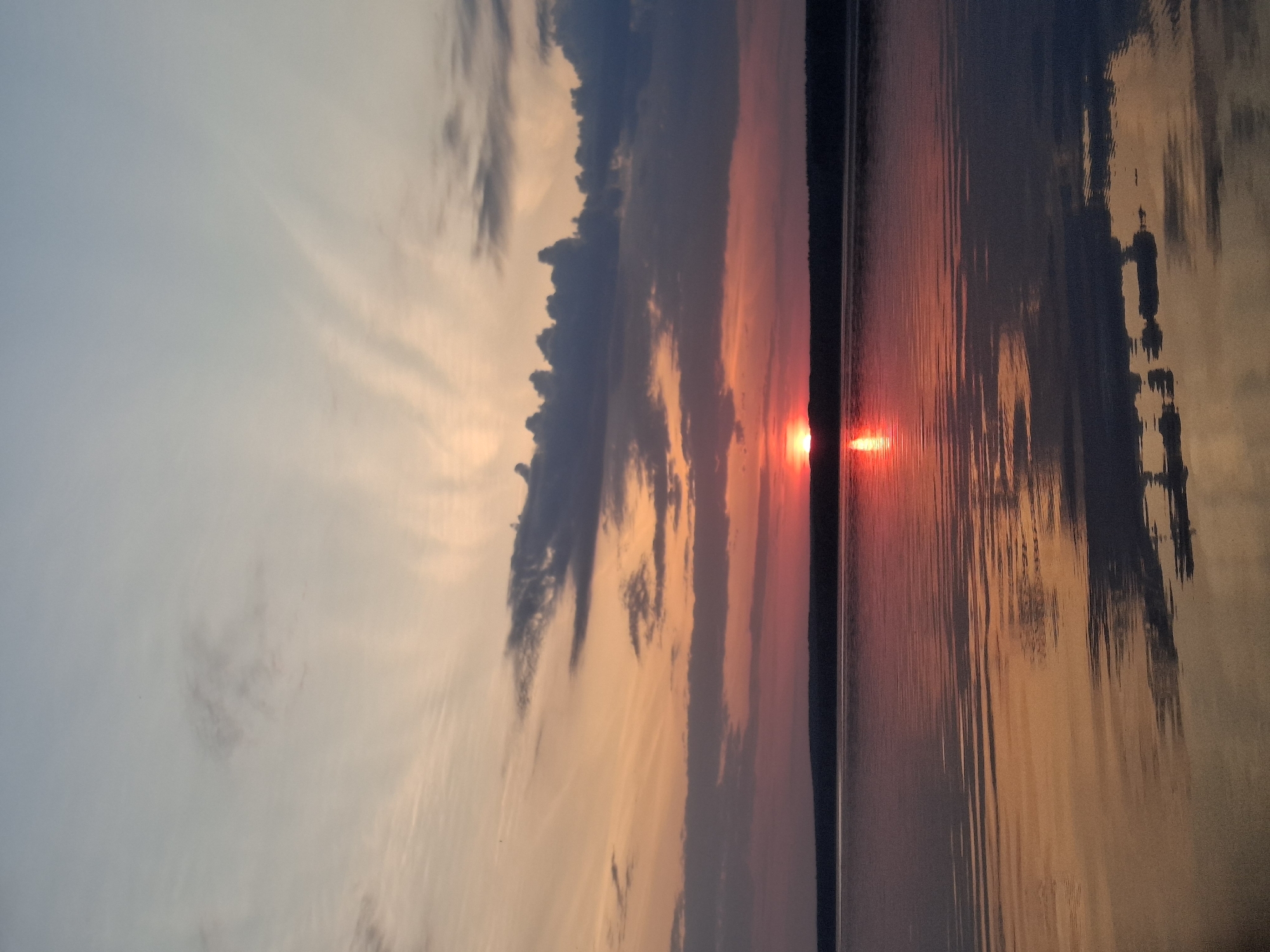 The image shows a sunset over water. The sun is positioned at the horizon line, reflecting a bright red-orange glow on the water's surface. The sky features a gradient of colors from light blue to orange and pink, with streaks of darker clouds. The scene appears to be near Chaikovsky, Russia. The composition includes the horizon, sky, water, and clouds. There is a landmass in the middle, creating a distinct division between the sky and water. Reflected silhouettes of structures or objects can be seen on the right side of the water's surface.