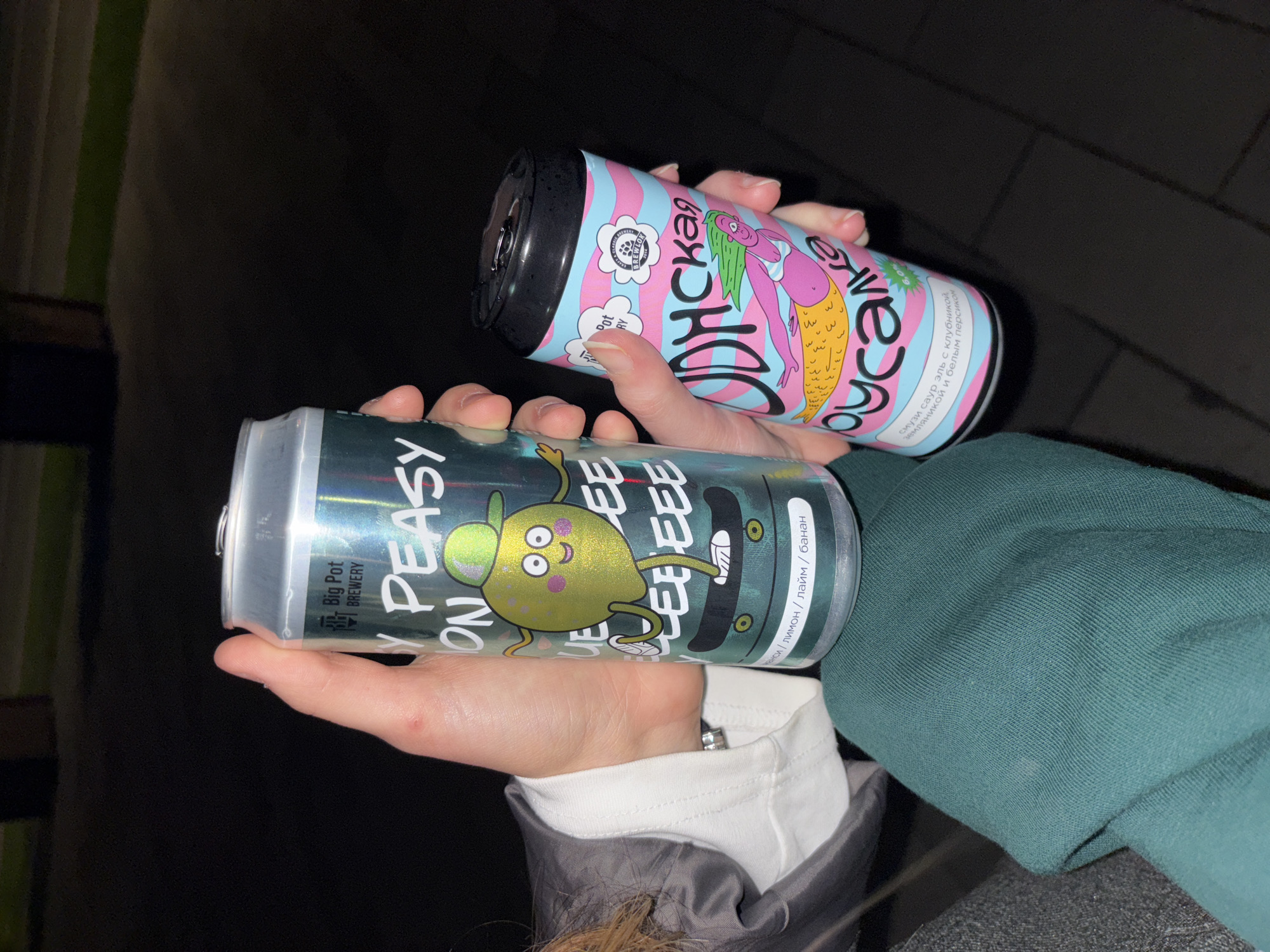 In a dimly lit outdoor setting, possibly at night, a person's hands are holding two cans of craft beer. The can on the left, labeled "Big Pot BREWERY," features a cartoon lime character wearing a baseball cap and riding a skateboard. The can on the right, also from "Big Pot BREWERY," has a pastel pink and blue design depicting a cartoon mermaid. The background suggests a paved area, possibly a sidewalk or plaza, with blurred out structures in the distance, implying an urban environment. The overall scene is casual and indicative of enjoying beverages outdoors. It is not possible to determine the specific location in the city of Perm, Russia, from the visual information provided.