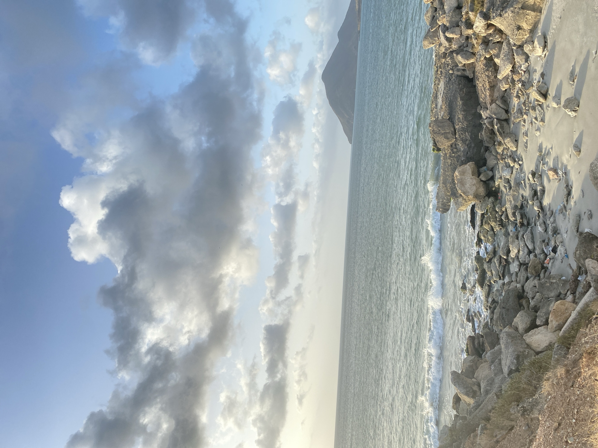 The image captures a coastal scene in Juan Griego, Venezuela, featuring a rocky shoreline meeting the ocean. A small mountain or hill protrudes on the horizon. The sky is filled with clouds, obscuring most of the blue. The sea appears calm with small waves washing onto the shore. The shoreline is rugged, composed of large rocks, sand, and some vegetation.