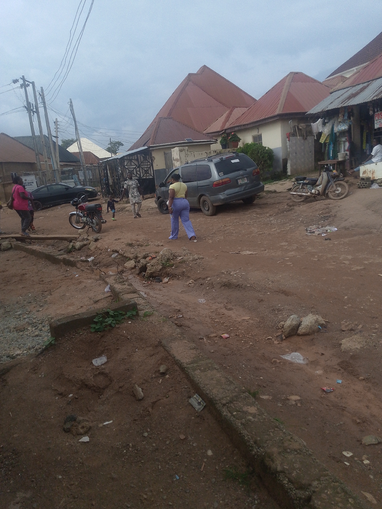 In Kurunduma, Nigeria, a dusty, unpaved street is depicted. A grey minivan is parked on the right side of the road. A woman wearing a yellow top and blue pants walks away from the camera. A motorcycle is parked near the minivan. On the left side of the street, a black sedan is visible. A woman in a pink top stands near the sedan. Several buildings with red roofs line the street. Power lines and utility poles are present overhead. The ground is covered in dirt and debris, with some evidence of erosion and puddles.