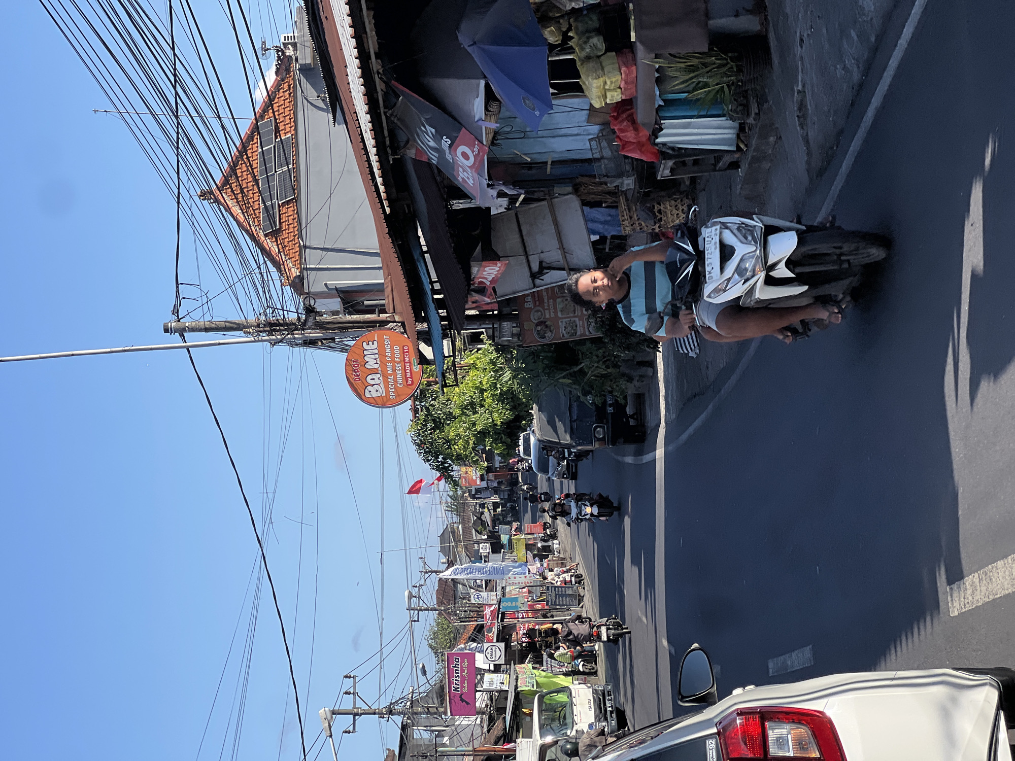 The image depicts a street scene in Denpasar, Indonesia, on a bright, sunny day. A busy road runs through the center of the frame, with a procession of motorcycles and a few cars traveling along it. On the right side of the road, there are several shops and buildings, some with corrugated iron roofs. One shop has a circular orange sign that reads "DEPOT BA MIE SPECIAL MIE PANGSIT CHINESE FOOD by MADE MCI 10". A person is riding a white scooter in the foreground, with another person sitting behind them. Both are wearing casual clothing. The background shows more buildings and dense overhead power lines, a common sight in urban Indonesian settings. The overall impression is of a bustling commercial street, characteristic of Denpasar.