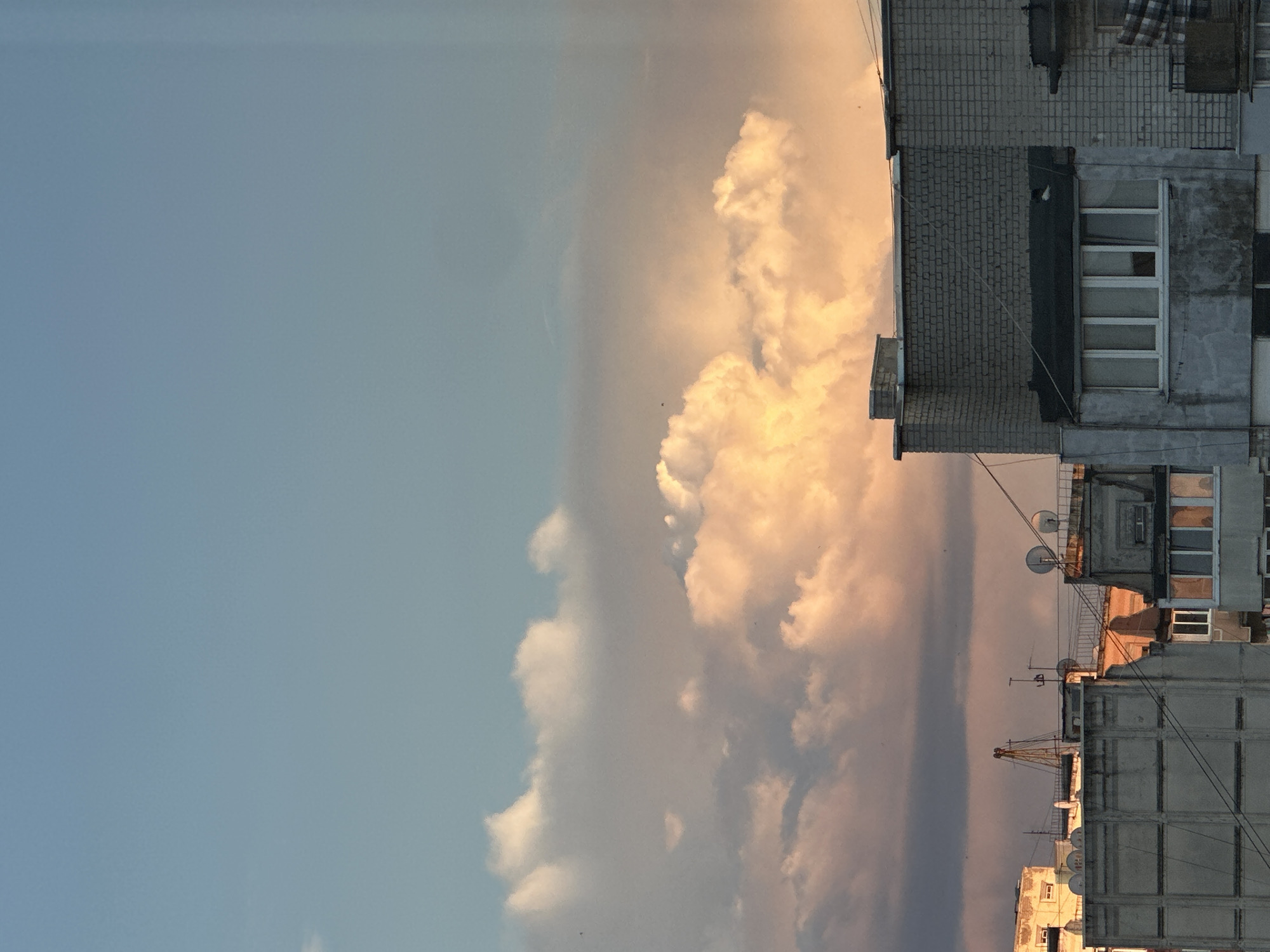 A twilight cityscape viewed from above shows a sky filled with dramatic clouds illuminated by the setting sun. The clouds range in color from soft whites and peaches to deeper grays, creating a textured canopy above the urban environment. Below, the rooftops of buildings are visible. These structures appear to be residential or office buildings, with varying architectural styles, some featuring visible satellite dishes and other rooftop equipment. The overall impression is one of urban life transitioning into evening, with the natural beauty of the sky providing a striking contrast to the man-made structures.