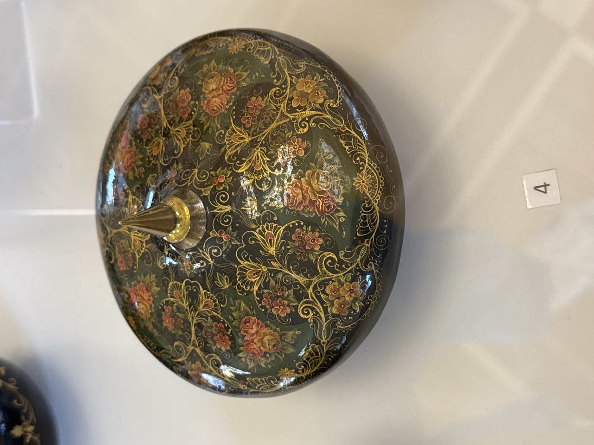 A domed, dark green lidded box, possibly made of wood and lacquered, sits on a white surface. The lid is intricately decorated with a floral and scroll pattern in gold, red, and orange. A golden, pointed finial rises from the center of the lid. In the lower right corner of the image, a small white square with the number "4" printed in black is visible on the white surface, possibly a label or marker. The background is a plain white surface with subtle shadows. The scene appears to be an exhibit or display, and given the context of the number "4" and the white surface, it may be part of a museum or gallery in Sochi, Russia, though there is no explicit visual evidence to confirm this location. There are no people or animals visible in the image.