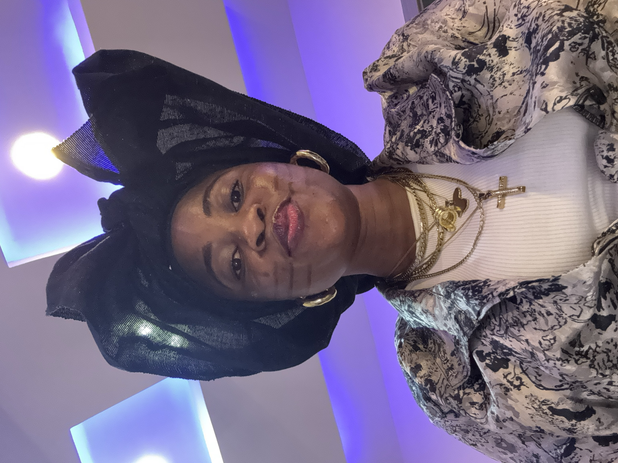 A portrait of a woman wearing a black headwrap, gold hoop earrings, and multiple gold necklaces, one with a cross pendant. She is also wearing a patterned jacket over a white top. The background features purple lighting and recessed ceiling lights. The woman is looking directly at the camera with her lips slightly pursed. The scene does not provide any information or clues about a location in Ajara, Nigeria.