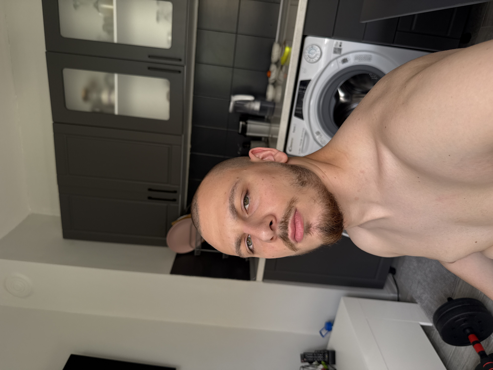 A shirtless man with a beard is in a kitchen. The kitchen has gray cabinets, black tiled backsplash, and a white washing machine. A dumbbell is on the floor. The location in the city of Stara Zagora, Bulgaria is not determinable from the image.