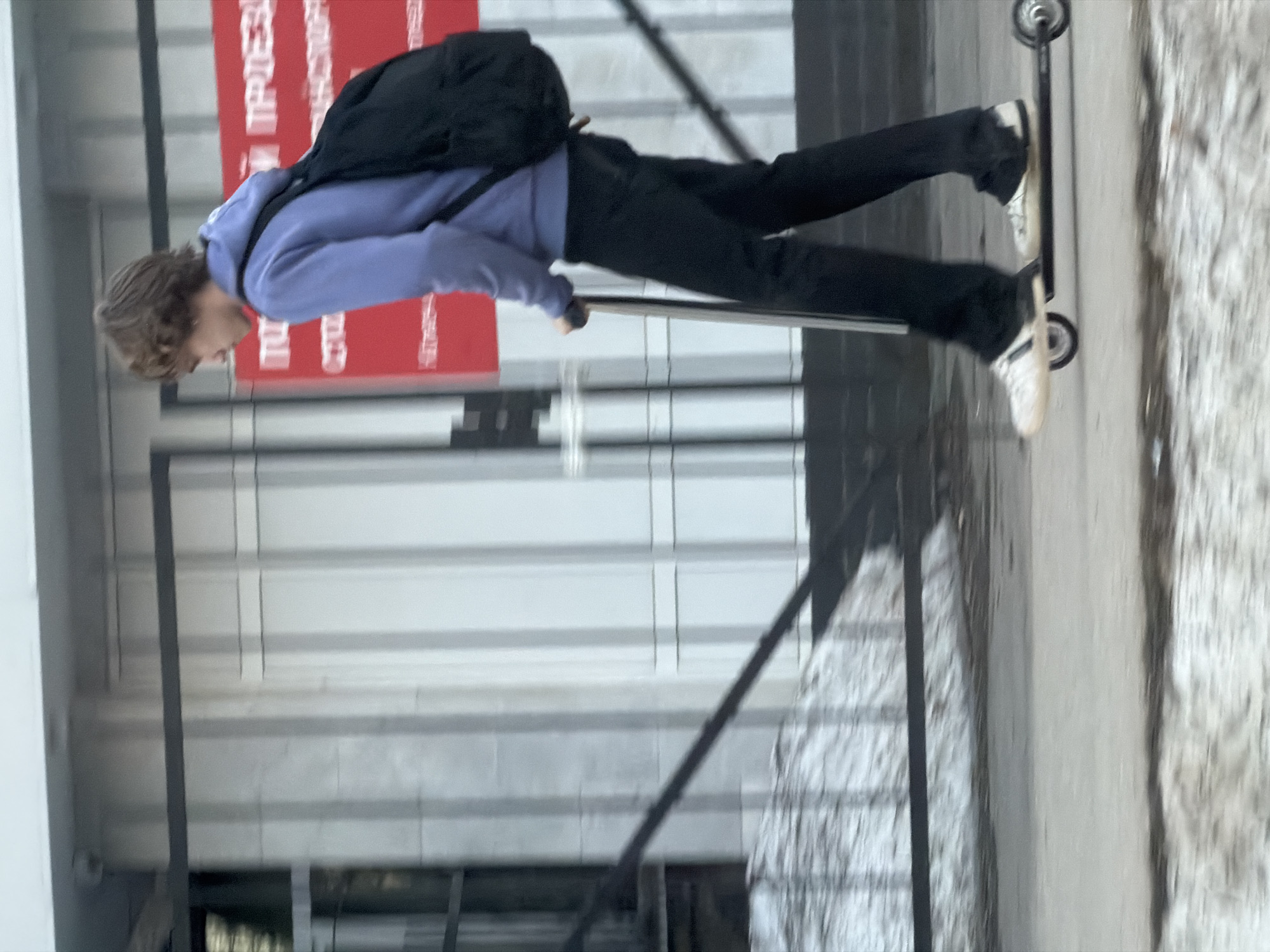 A person with a backpack is riding a scooter in Perm, Russia. The person is wearing a purple hoodie, black pants, and white shoes. They are riding on a sidewalk next to a building with a red sign. The background is blurred.
