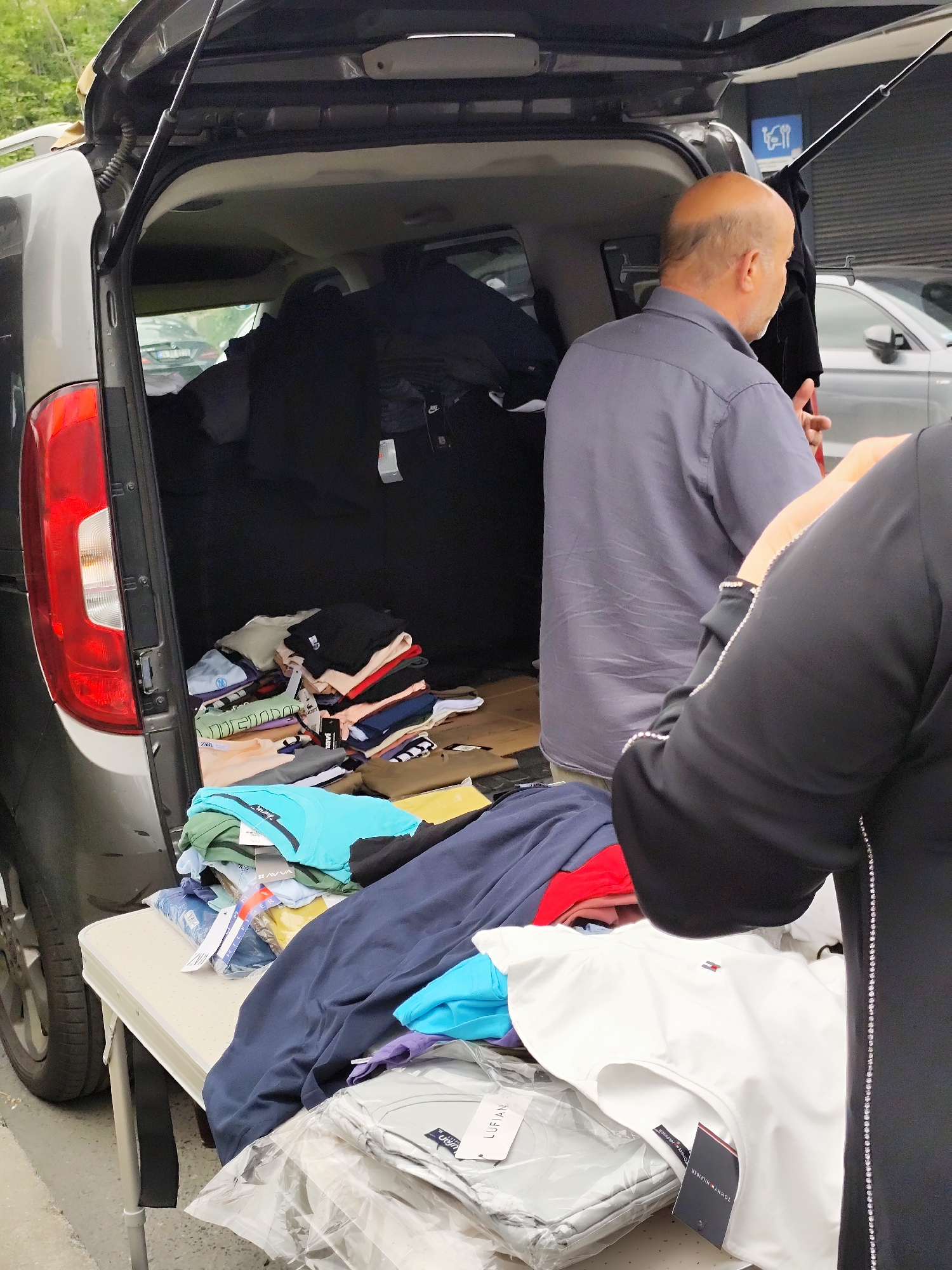 In an outdoor location in an unknown city, a car with its trunk open displays a large quantity of clothing. A bald man in a grey shirt stands near the open trunk, while another person, identifiable by their black clothing with rhinestone detailing, stands in the foreground. A white table holds more clothing items. The trunk of the car is packed with clothes, and the surrounding environment includes a paved surface and building in the background.