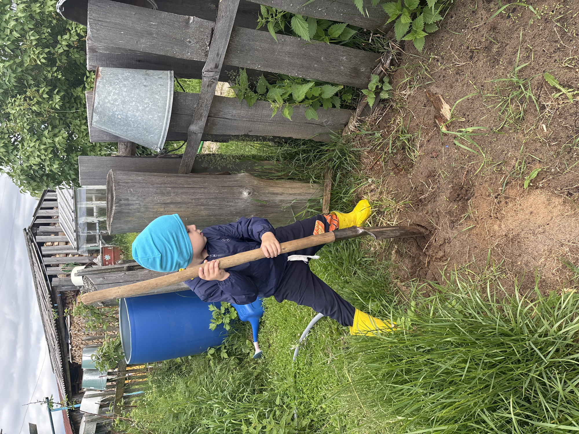 In a rural outdoor setting, a young child wearing a blue beanie, navy jacket, black sweatpants, and yellow rubber boots is digging in the dirt with a shovel. The child is positioned in the foreground, facing towards the right side of the frame, with the shovel embedded in the soil. The environment appears to be a backyard or garden area, with lush green grass, a wooden fence, a large blue barrel, and a dilapidated wooden structure in the background. The sky is overcast, suggesting cloudy weather. The location is identified as Kurgan, Russia.