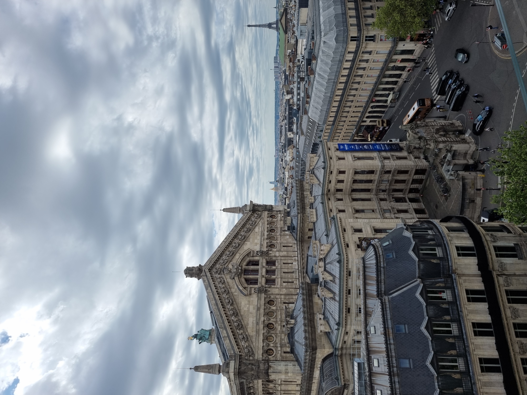 The image depicts a view over Paris, France, with a prominent ornate building complex in the foreground. In the background, the Eiffel Tower is visible amidst a cloudy sky. Various Parisian streets are visible, filled with vehicles and pedestrians. The sky is overcast, with dense cloud cover.