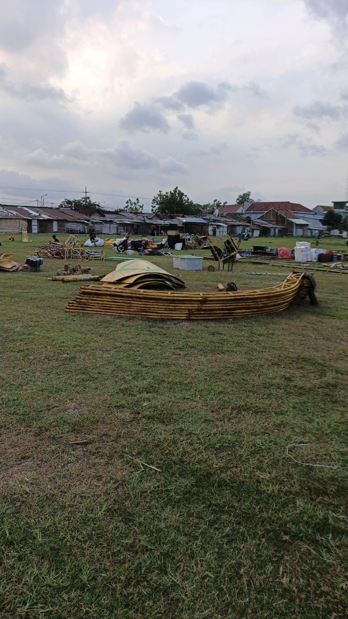 The image captures an outdoor scene in the city of Boyolangu, Indonesia, likely in a park or open field. The sky above is filled with clouds, suggesting either an overcast day or approaching dusk. In the foreground, a large, curved structure made of what appears to be bamboo poles, painted yellow, lies on the grass. The structure is segmented, with many layers of poles stacked on top of each other. Further back, various other objects are scattered across the grassy area, including what looks like playground equipment, a scooter, and what might be event props. In the background, a row of buildings with corrugated iron roofs and some houses are visible, indicating a residential or semi-urban area. A few individuals are present in the midground, some near the scattered objects and others obscured by them. The overall impression is of a location where an event has recently concluded or is in the process of being set up or dismantled.