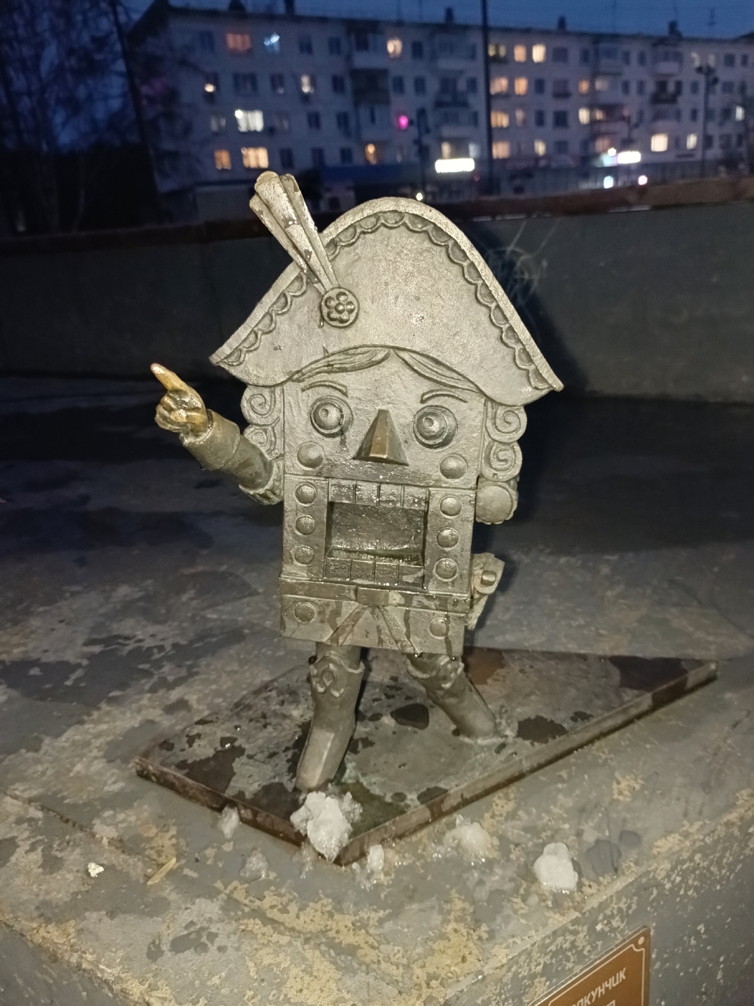A low-angle shot shows a metal sculpture of a nutcracker in the city of Chaikovsky, Russia. The nutcracker stands on a triangular metal base placed on a weathered, concrete-like surface. Snowflakes are scattered around the base. The nutcracker figure is detailed with a square face, decorative eyes, and a tricorn hat with a feather. One arm is raised with the index finger pointing to the right. In the background, an apartment building with illuminated windows is visible in the dark. The sky is dark blue.