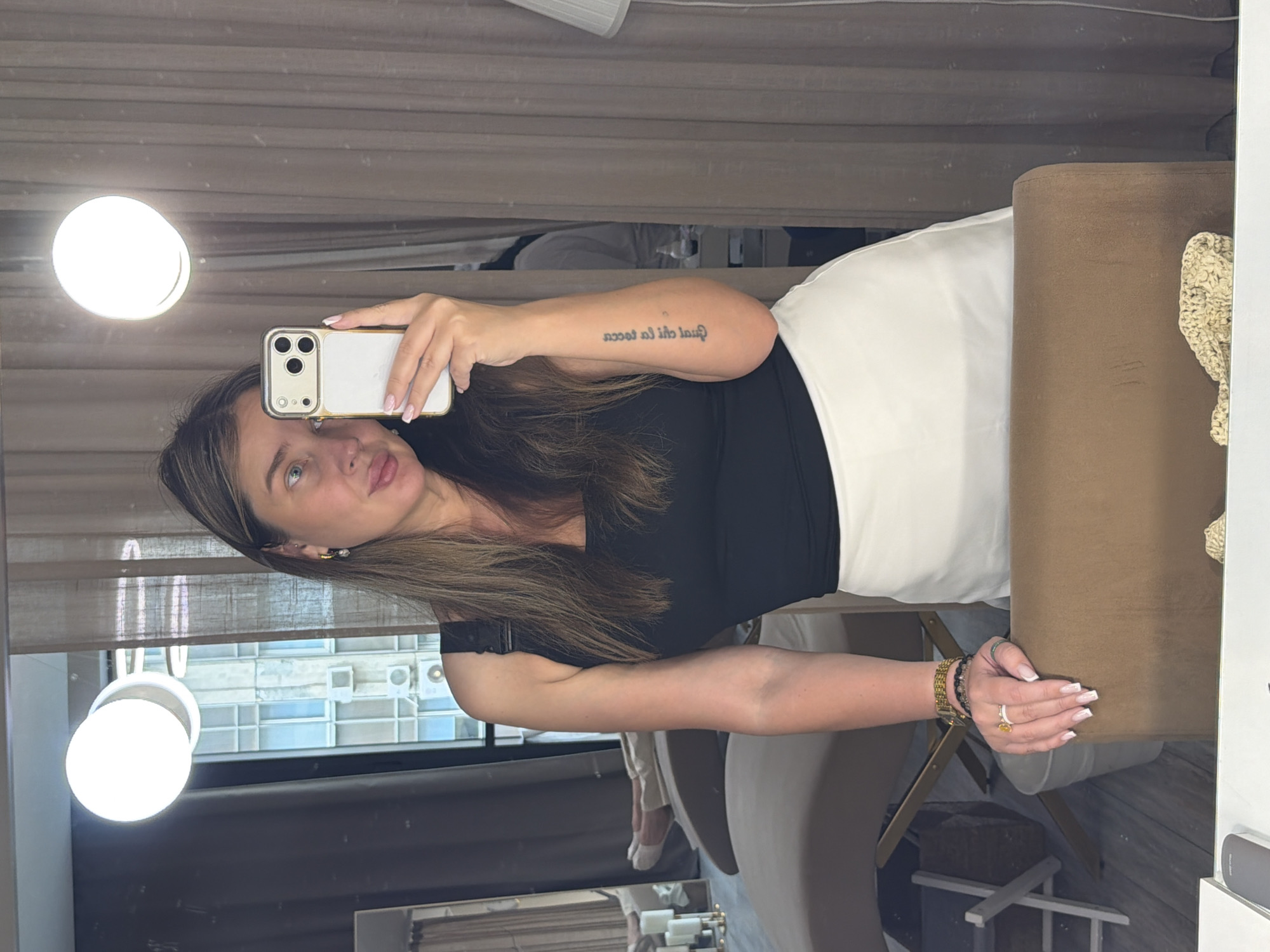 A woman with long brown hair is taking a selfie in a room with a large mirror. She is wearing a black tank top and a white skirt. She has a tattoo on her left forearm. Her right hand is holding a smartphone. She is wearing jewelry on her left hand, including a ring with a yellow stone and bracelets on her wrist. In the background, there are curtains, a lamp, a massage table, and a view of a building with windows. The mirror reflects a portion of the room.