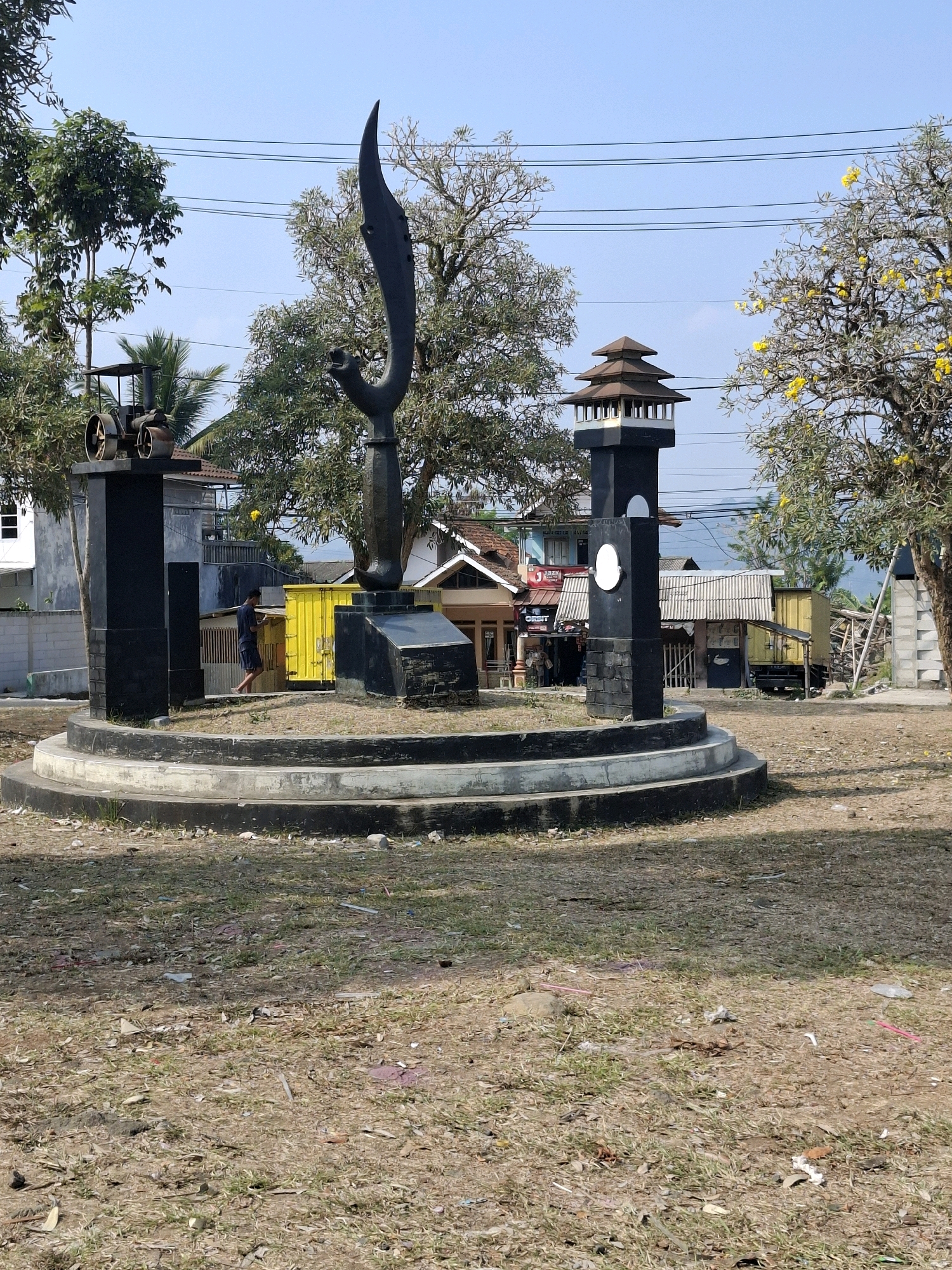 The image depicts an outdoor scene in Sukabumi, Indonesia, featuring a monument with a dark, abstract sculpture resembling a bird's head and beak, set against a clear blue sky. The monument is surrounded by a circular, tiered base. To the left of the main sculpture, a black pedestal supports a miniature steam engine model. To the right, another black pedestal features a traditional tiered roof structure, resembling a pagoda, and a circular white emblem. The ground surrounding the monument is dry and covered with sparse grass, fallen leaves, and scattered debris. In the background, several buildings with corrugated metal roofs are visible, along with trees, including some with yellow flowers. A young person wearing shorts and a t-shirt is walking in the mid-ground, near a bright yellow shipping container. Power lines crisscross the upper portion of the image.