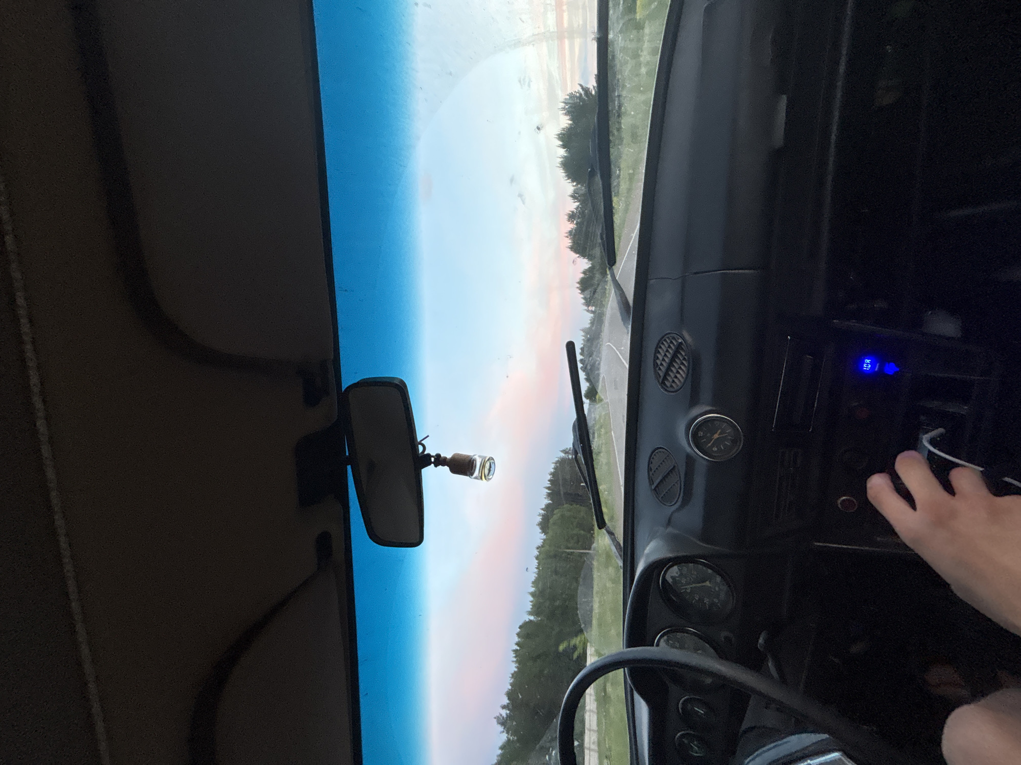 Captured from inside a car, the image presents a driver's perspective on a street view in Прикамский, Russia. The car's interior, including the dashboard, steering wheel, rearview mirror, and a hand, dominates the foreground. Through the windshield, the outside reveals a road flanked by trees. The sky is a mix of pale blue and pink, possibly indicating dawn or dusk.