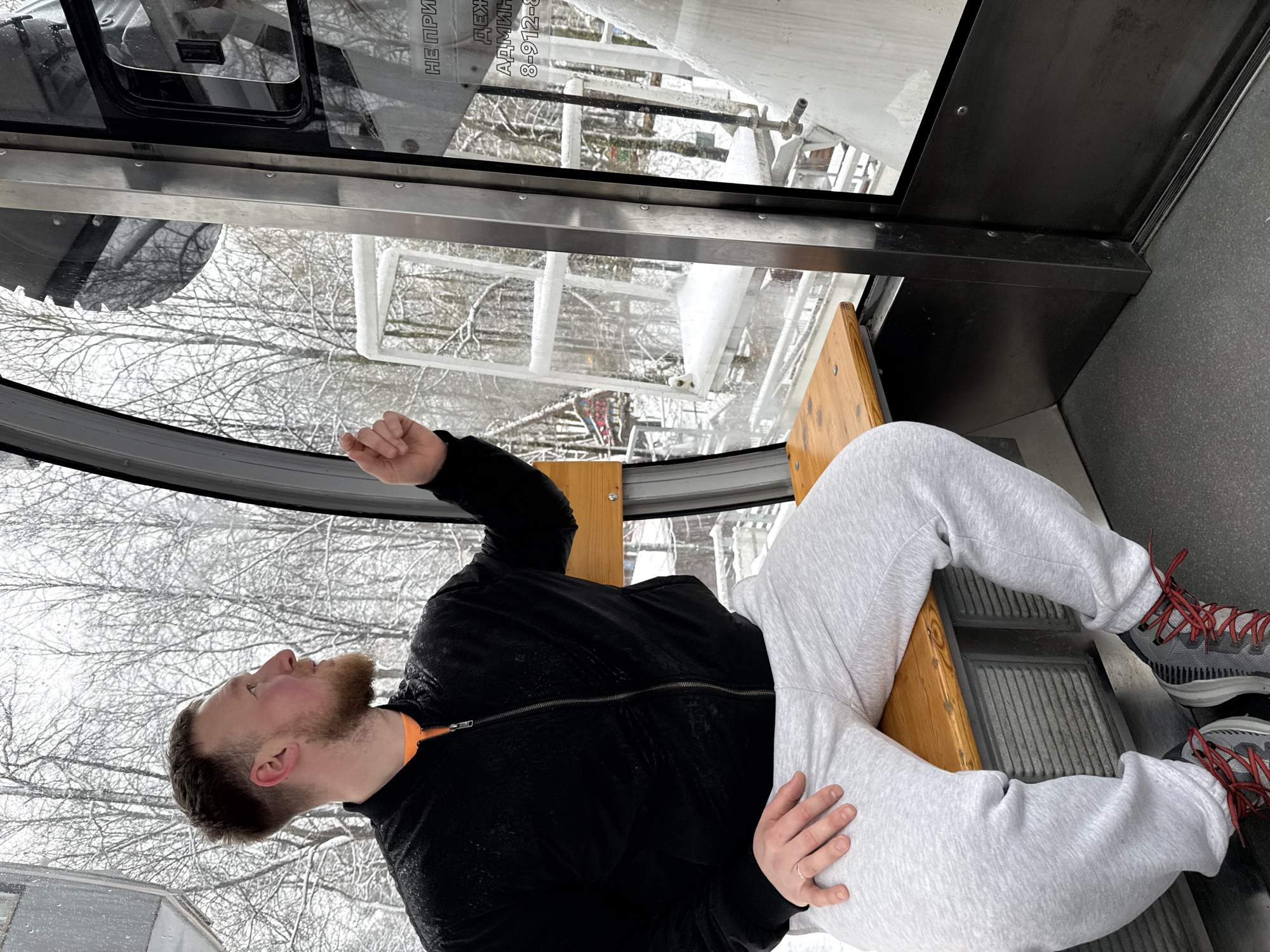 A man is sitting inside a glass-walled structure with a view of snow-covered trees and buildings. The man is wearing grey sweatpants, a black jacket with an orange collar, and grey sneakers with red laces. He is seated on a wooden bench. Through the glass, snow-laden branches and structures are visible. A partial view of lettering in a language other than English is on the glass. Given the location stated in the prompt, the view through the window likely depicts a winter scene in Perm, Russia. The structure appears to be a gondola or similar enclosed cabin.