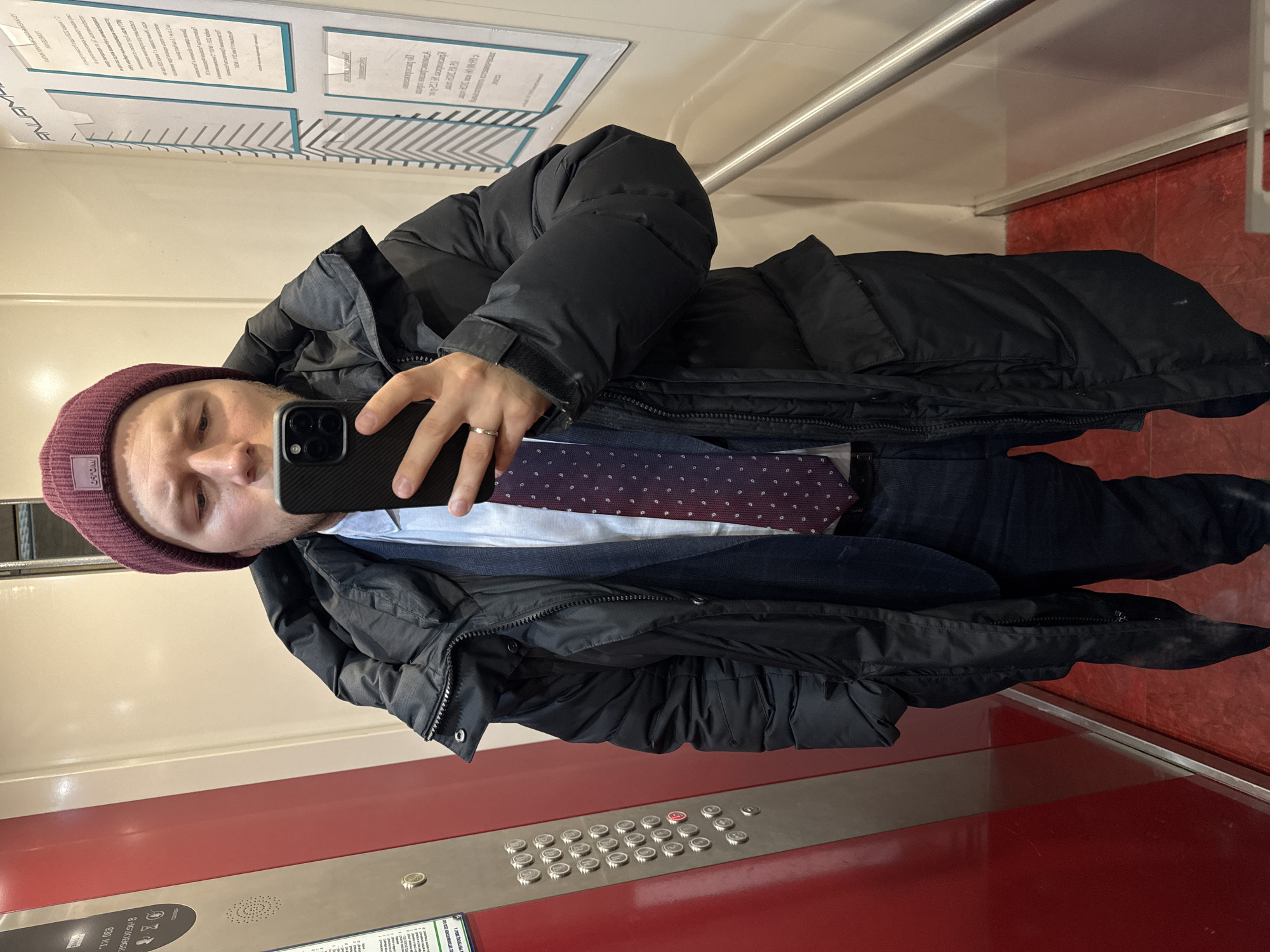 A man in a maroon beanie and a black puffy jacket takes a selfie in an elevator with a red and white interior, and a panel of silver elevator buttons, in Krasnoyarsk, Russia. He is wearing a light blue shirt and a dark blue tie with a white pattern. His phone is black. There are signs posted on the elevator wall.