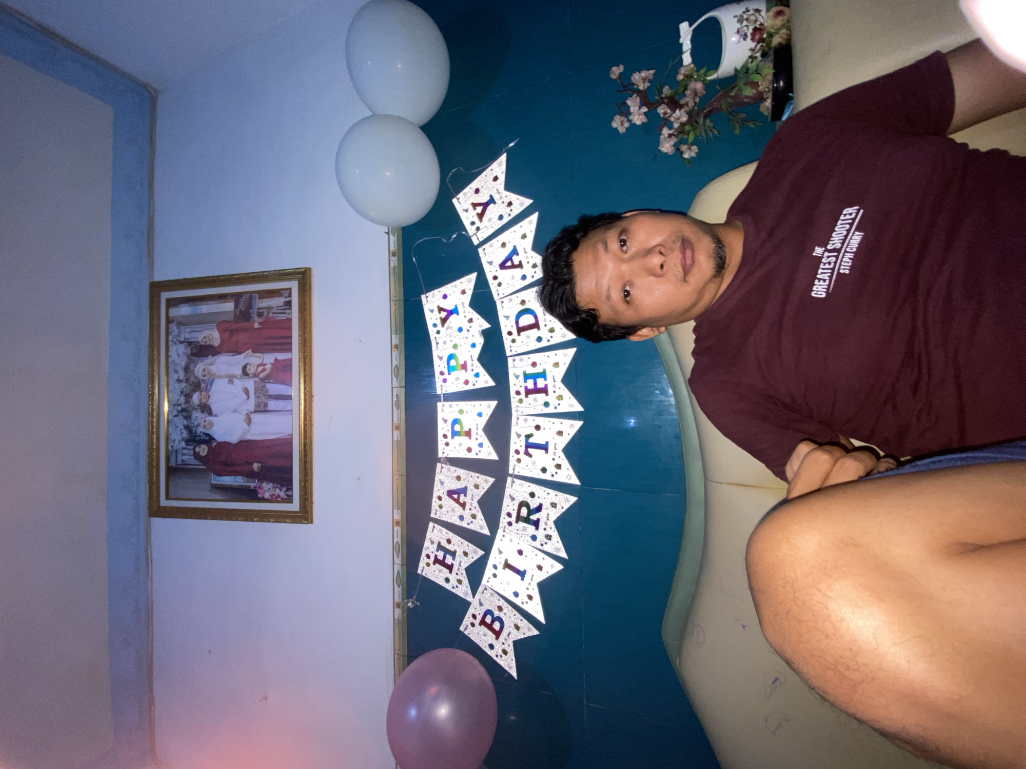 The scene appears to be an interior decorated for a birthday celebration. A middle-aged Asian man wearing a maroon t-shirt with the text "THE GREATEST SHOOTER STEPH CURRY" is reclining on a cream-colored sofa. A "HAPPY BIRTHDAY" banner hangs on a dark blue wall, flanked by two white balloons and a single pink balloon. A framed photo, possibly a family portrait, is mounted on the wall to the left. A vase with flowers is positioned above the sofa. The location is inferred to be Gelam, Indonesia.